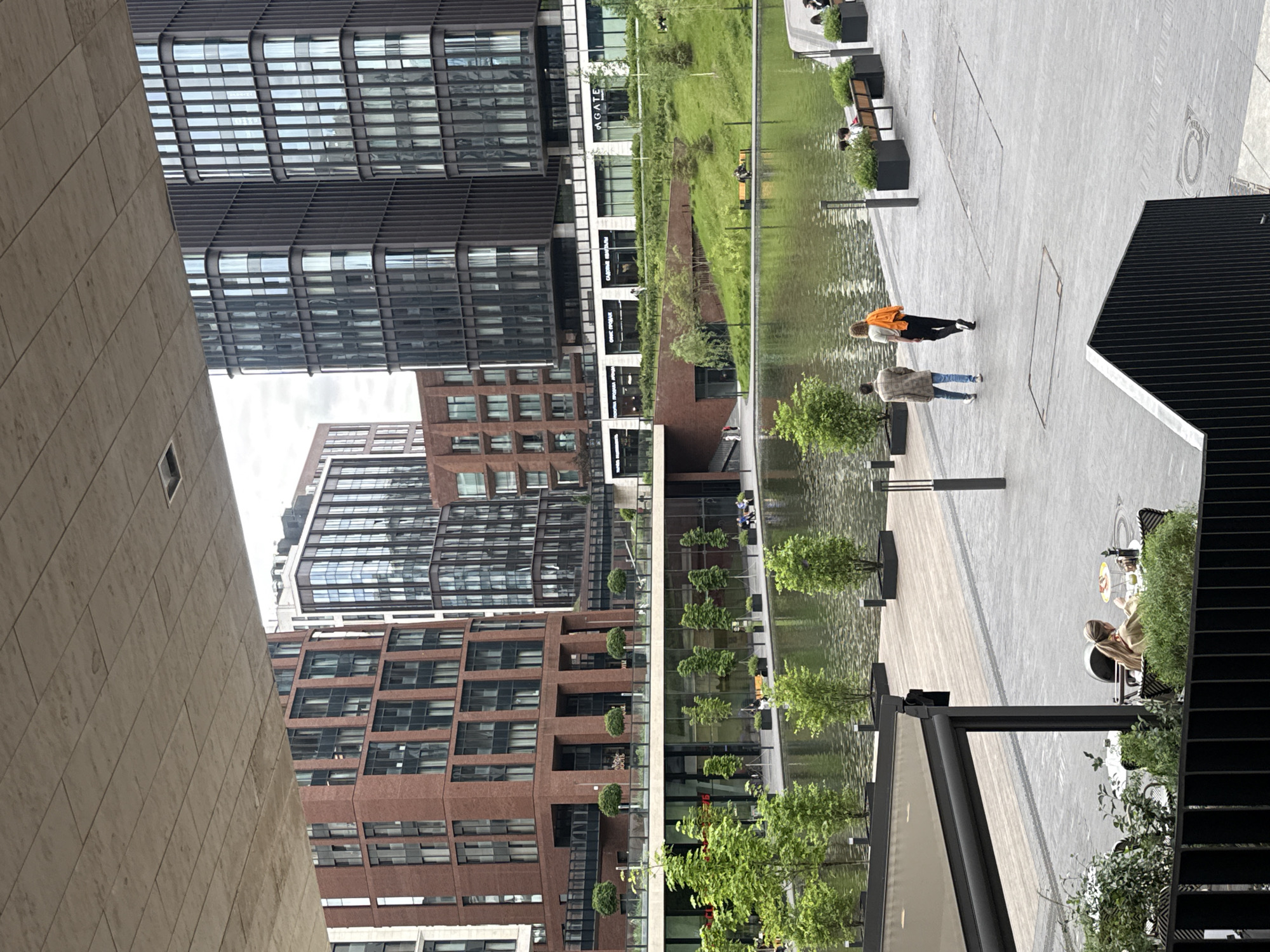 The image captures an outdoor urban complex in Moscow, Russia, characterized by modern architecture and a landscaped public space. The scene is dominated by several multi-story buildings of varying designs. On the left, there are tall structures featuring dark, reflective glass facades and dark metal paneling, interspersed with brick buildings showcasing a grid of windows.
Running through the middle of the complex is a long, narrow water feature or canal, its surface reflecting the surrounding buildings and sky. This water body is bordered by meticulously maintained green landscaping, including small, neatly trimmed trees and low bushes.
To the right of the water feature is a spacious, paved pedestrian area. Several people are visible, engaged in various leisurely activities. Two individuals are walking along the pathway: one person wears a bright orange top and black pants, while another wears a plaid jacket over blue jeans. Further along the path, closer to the foreground, a person with long hair is seated at an outdoor table with a yellow plate, appearing to be dining or having a drink. In the background, other individuals are seated on benches near the water, suggesting a place for relaxation and socializing.
The time of day appears to be daytime, with soft, diffused lighting suggesting an overcast or cloudy sky. The overall atmosphere is calm and inviting, depicting a functional yet aesthetically pleasing urban environment. Visible text on one of the commercial building facades includes "AGATE" in Latin script, and several signs in Cyrillic, such as "САЛОН КРАСОТЫ" (Beauty Salon), "ОПТИКА" (Optics), "СУПЕРМАРКЕТ" (Supermarket), and "АПТЕКА" (Pharmacy), indicating various businesses operating within the complex.