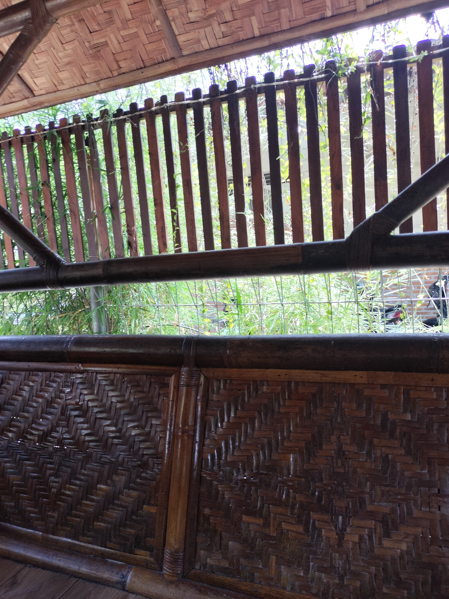 The image shows a close-up view of a bamboo structure, likely part of an outdoor seating area or pavilion. The foreground features a woven bamboo bench or wall with a detailed pattern. Above this, a horizontal bamboo railing runs across the frame. Beyond the railing, a fence made of vertical wooden planks is visible, with lush green foliage and vines growing through and over it. Sunlight filters through the fence and greenery, creating a dappled light effect. In the background, out of focus, are hints of a building with brickwork and possibly some vehicles. The overall impression is one of a rustic, natural setting. The location is specified as Tambonbaroh, Indonesia.