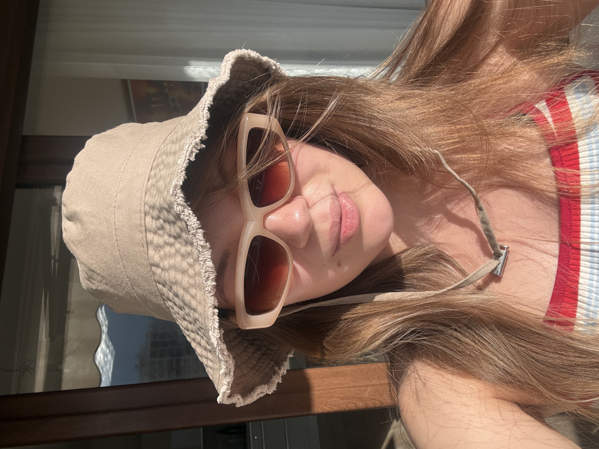 A young individual, appearing female, is depicted in a sunlit, close-up photograph, facing directly towards the viewer. The subject has long, light brown hair and wears a light beige bucket hat with a distressed, frayed brim. Light-framed, rectangular sunglasses with brown gradient lenses cover the eyes. A small mole is present on the philtrum above the upper lip. The subject is wearing a striped garment featuring prominent red, white, and light blue horizontal bands. A narrow, light-colored fabric strap, possibly from the hat, with a small metallic rectangular slider, rests on the subject's chest. Intense sunlight originates from the right, strongly illuminating the right side of the subject's face and hat, and casting subtle shadows on the left.
The background shows an interior space with a large window or glass door. Through this opening, a distant outdoor environment is visible. The outdoor scene features a body of water exhibiting distinct wave patterns, indicating a coastal location. Beyond the water, several multi-story buildings, consistent with modern residential or hotel architecture, line a distant shoreline. These structures are backed by mountainous or hilly terrain under a bright sky. The combination of coastal waters, distant architecture, and mountains strongly suggests a location within the city of Budva, Montenegro. Dark window frames and light-colored interior curtains are also discernible in the background.