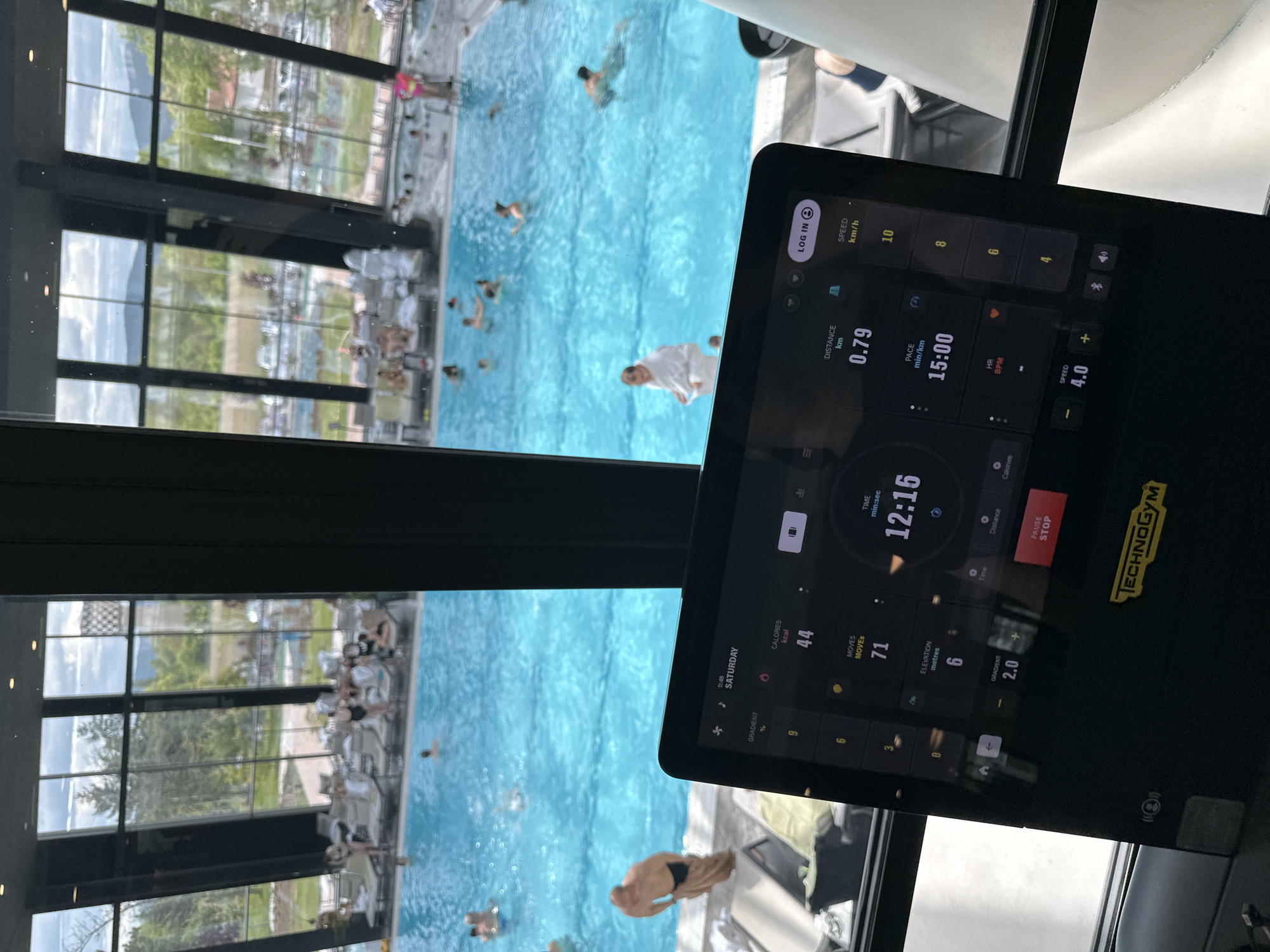 The image shows a person using a Technogym treadmill in what appears to be a fitness center or spa, with a view overlooking an outdoor swimming pool. The treadmill screen displays statistics like distance (0.79 km), pace (15:00 min/km), time (12:16), calories (44 kcal), moves (71) and speed (4.0 km/h). Through the large windows, people can be seen swimming in the pool, and sunbathing on deckchairs. The location is likely a resort or hotel with wellness facilities. I have no basis for concluding this image depicts the city of Kazanlak, Bulgaria.