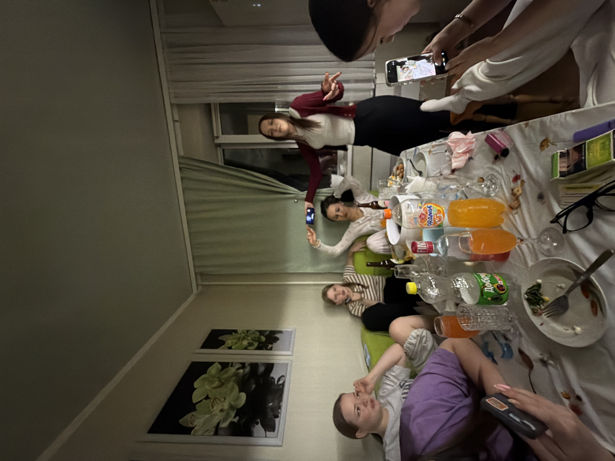 This image captures a casual indoor gathering of at least six young women, presented with a 90-degree clockwise rotation. The scene appears to be a living room or common area in Perm, Russia, as suggested by the Russian brand "ДОБРЫЙ" (Dobry) visible on a beverage bottle.
The women are engaged in various relaxed activities around a table and on a green couch. One woman, wearing a white top, maroon cardigan, and black skirt, stands and strikes a pose with her hands. Another woman in a light top holds a smartphone, seemingly taking a picture of the posing individual. A third woman in a striped top sits on the green couch, looking towards the camera. Another person is partially visible in purple shorts or pants, holding a remote control or a similar device. Several other individuals are present, with at least two more also holding smartphones, indicating that capturing photos or videos is part of the social interaction.
The table is covered with a patterned tablecloth and holds an assortment of items: empty and partially filled plastic bottles (some with orange-colored drinks, one with green liquid), glasses, plates with food remnants (including salad), napkins, and eyeglasses. The brand "ORANGE" is also visible on one of the bottles.
The setting includes light-colored walls, green curtains, and a window with horizontal blinds in the background. Two framed pictures featuring green orchids are displayed on one wall. The interior lighting suggests it is likely evening or night. The overall atmosphere is one of a relaxed social gathering among friends.