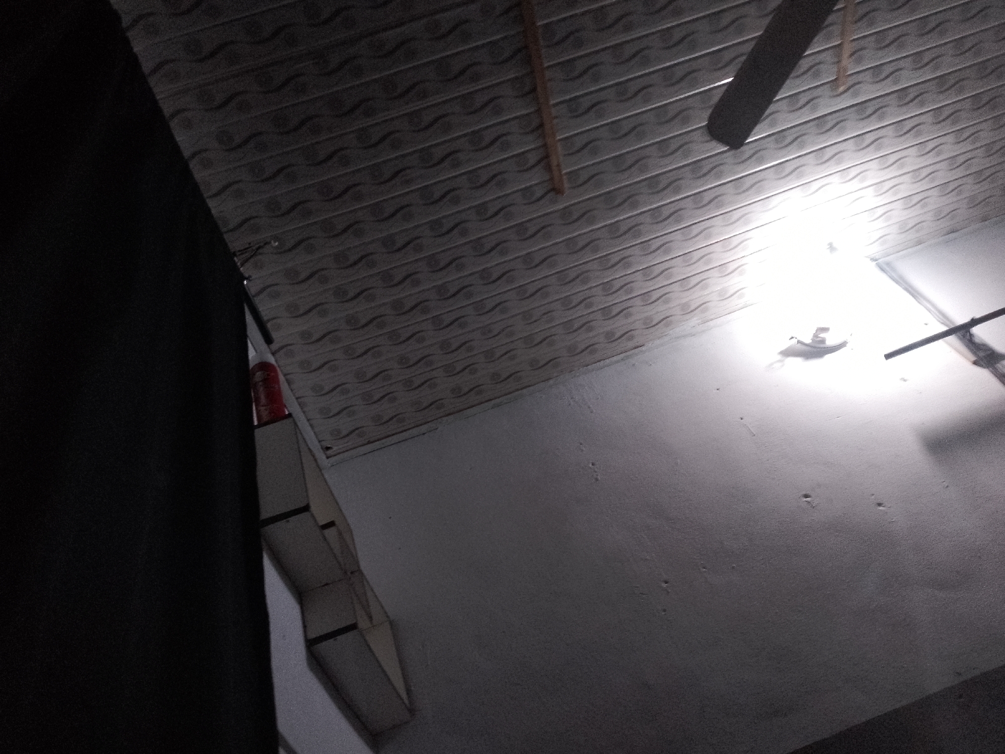 The image shows a low-angle view of a ceiling in Jauro Yinu, Nigeria. The ceiling appears to be made of textured panels with a wavy pattern. A ceiling fan with dark blades is partially visible at the top left of the image, and a single light bulb is illuminating the scene from the right. A dark, opaque object is on the left side of the frame, obscuring a portion of the ceiling and wall. Below the ceiling, the top portion of a white wall is visible, with a few small imperfections or marks. In the lower left quadrant, part of a structure with a stepped, box-like design is visible, with some dark trim. This structure appears to be made of light-colored material, possibly cardboard or wood, and may be decorative or functional. The lighting is bright around the bulb, creating a significant glare, while the rest of the scene is darker.