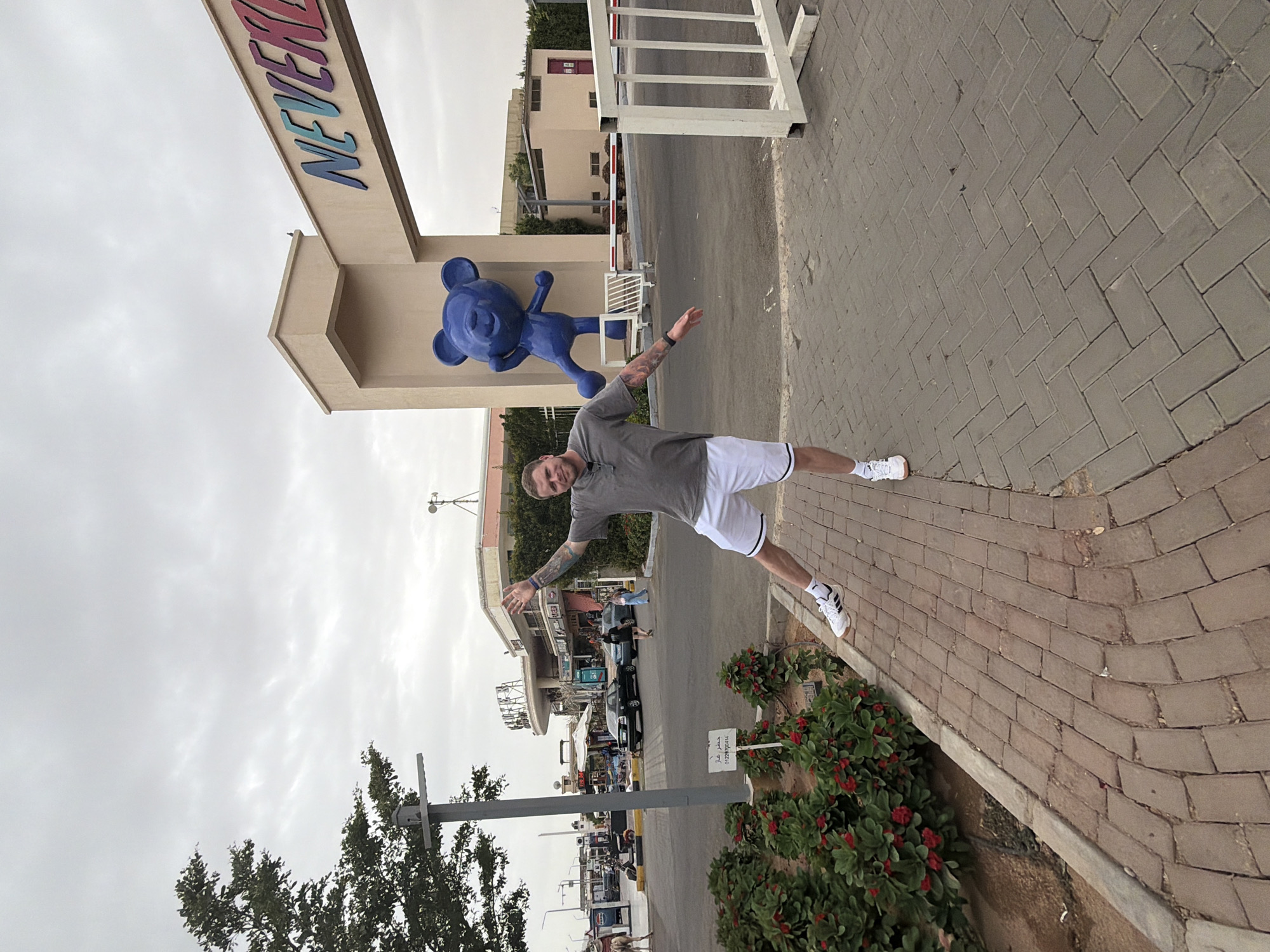 A man poses with outstretched arms in front of the Neverland gate in Sharm El Sheikh, Egypt. He is wearing a gray t-shirt, white shorts, and Adidas sneakers. The gate features a blue Mickey Mouse statue. The background includes a cloudy sky, buildings, cars, and streetlights.