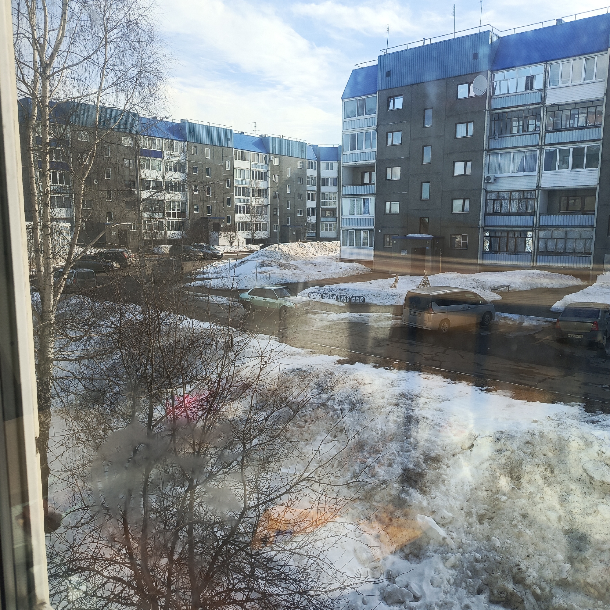 The image depicts an exterior urban scene in the city of Марковский, Russia, viewed from an elevated window, likely during late winter or early spring. The foreground shows the bare branches of deciduous trees partially obscuring the view, with significant, melting snow covering the ground. Reflections are visible on the windowpane, including internal elements and bright light flares.
In the midground, a wet asphalt road or parking area winds through the scene, bordered by large, dirty snow piles. Several vehicles are present: a light green sedan (possibly a Lada) is parked facing left, a silver minivan (possibly a Nissan Serena) is parked further right, and a dark sedan (possibly a Renault Logan) is parked behind the minivan. Blue metal bicycle racks and a bench are visible near the snow piles.
The background is dominated by a complex of multi-story, grey concrete apartment buildings, characteristic of Soviet-era architecture. These buildings feature numerous windows and enclosed balconies, with distinctive blue corrugated metal facades on their upper sections and roofs. Antennas are visible on some rooftops, and one building displays a satellite dish and an air conditioning unit. The sky above is partly cloudy with patches of clear blue. No individuals or specific actions are discernible within the frame.