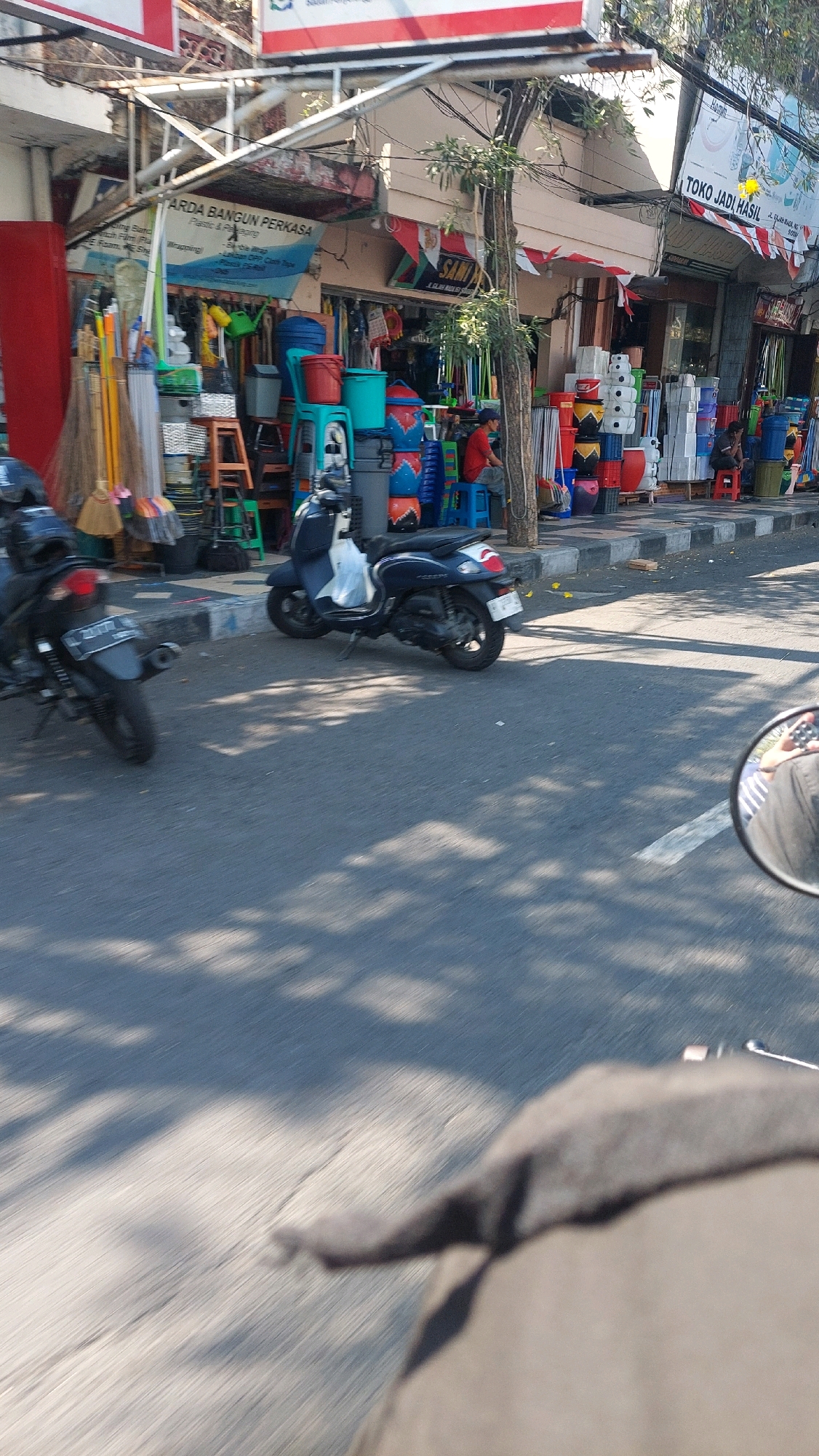 This is a street-level, eye-level, vertical, medium shot of a street scene in Sidoarjo, Indonesia, likely in the afternoon due to the shadows. On the left, a dark motorcycle is parked at an angle, facing away from the camera. To the right of it, a dark blue scooter is parked. Both motorcycles are parked on the sidewalk adjacent to a shop. The shop displays various household items including buckets, chairs, and brooms. Some of these items are stacked on the sidewalk. There are a few people visible. On the far right, a person is seated on a blue stool outside the shop. Another person is partially visible behind the stacked items on the right. The street is paved and shows some cracks. Shadows from trees are cast across the street and sidewalk. A white line is visible on the street. The mirror of a motorcycle, from which the photo is taken, is visible in the bottom right corner. The shop's signage is visible, including "ARDA BANGUN PERKASA Plastic & Packaging" and "TOKO JADI HASIL".