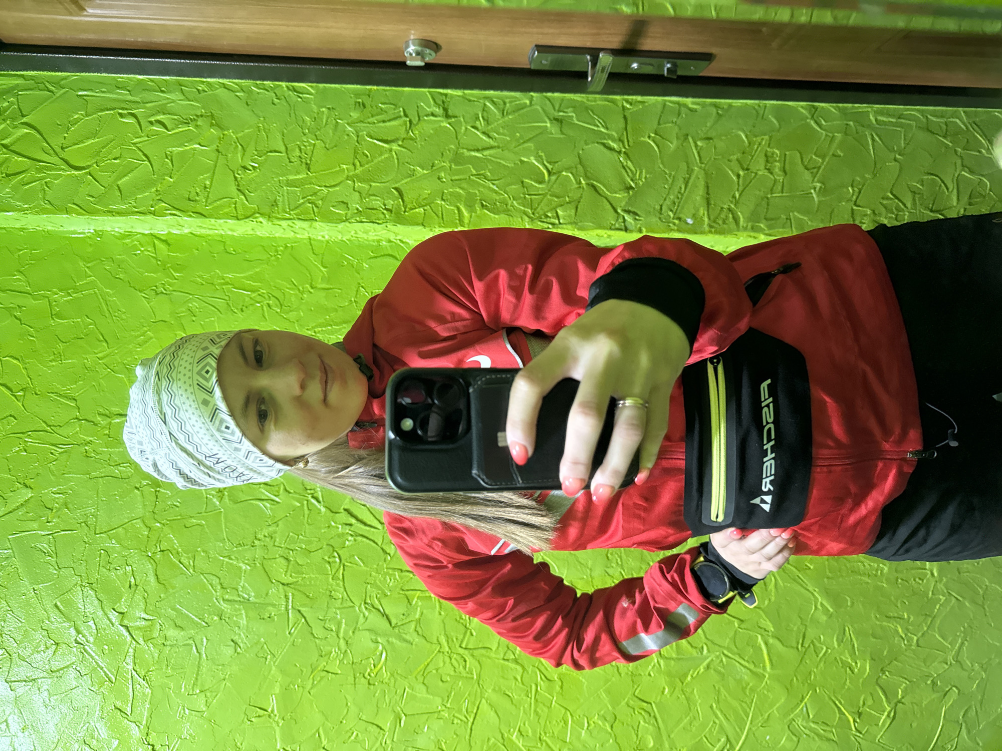 A woman is taking a selfie indoors against a vibrant green textured wall. The upper portion of a wooden door frame, secured with a simple latch, is visible at the top. The woman is positioned toward the left of the frame, wearing a white patterned headscarf, a red jacket with reflective stripes on the sleeves, and black bottoms. She holds a black smartphone in her right hand, the screen facing away from the camera, and is wearing a ring on her finger. A black and yellow fanny pack with the word "FISCHER" in reverse lettering is slung across her chest. A black wristwatch is visible on her left wrist. The photograph appears to have been taken in Chaikovsky, Russia, as the woman has identified herself as being located there in prior questions.
