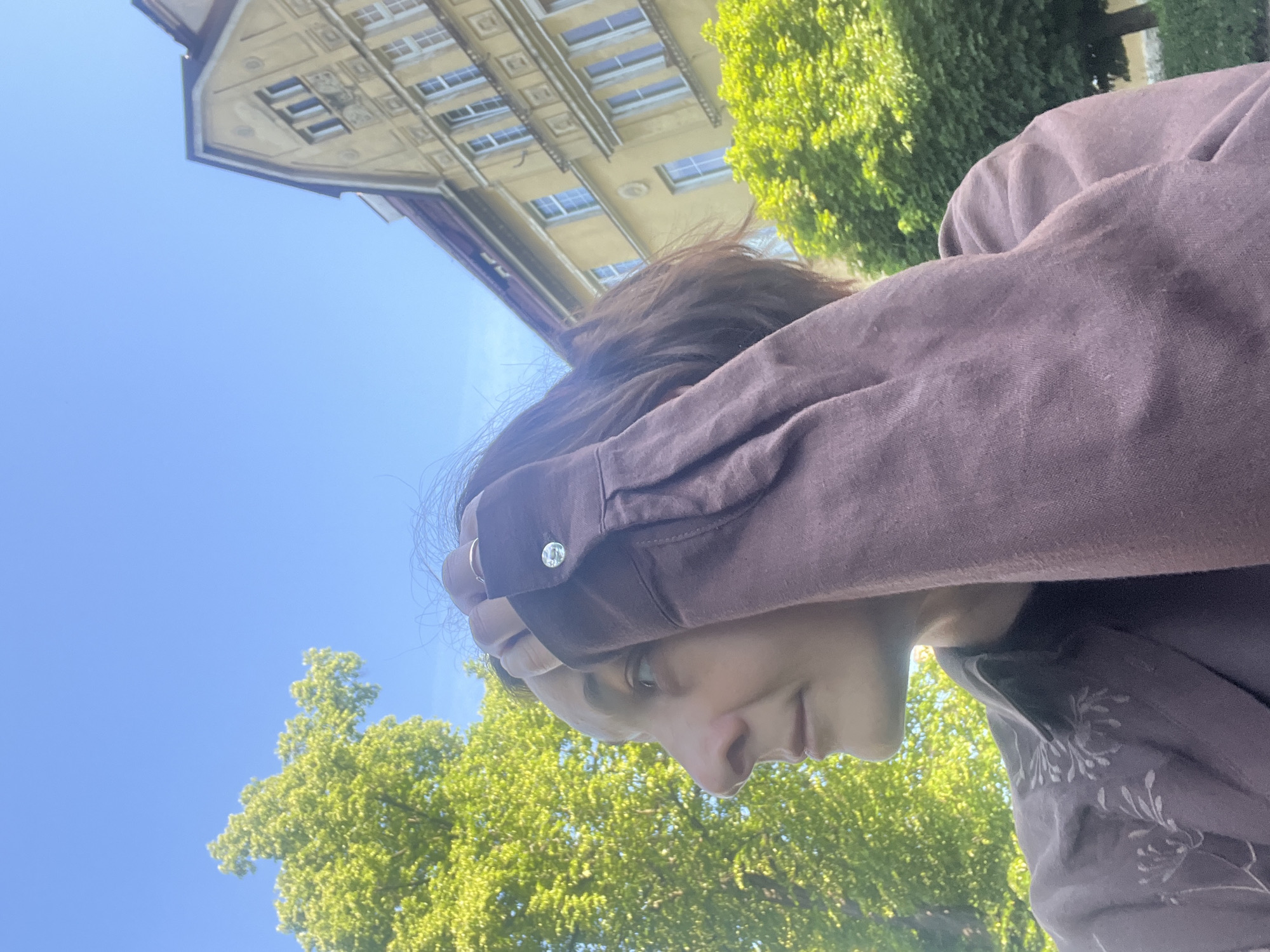 A low-angle, medium shot captures a person outdoors in front of a yellow building with a red-tiled roof. The person, shown from the side and slightly from the back, has short, dark hair and is wearing a dark brown shirt with a light-colored floral pattern on the collar. They are holding their right arm up to their head, with their hand touching their hair. A ring is visible on their right index finger. The sky is clear and blue, with bright green foliage from a tree on the left side of the frame and another tree on the right. The building features multiple windows and architectural details.