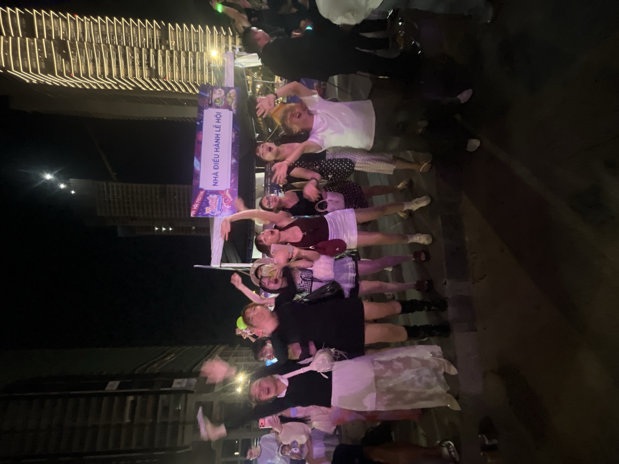 The image captures a nighttime scene in Da Nang, Vietnam, likely during a festival. A group of young people, predominantly women, are enthusiastically celebrating. They are positioned in front of a large banner that reads "NHÀ ĐIỀU HÀNH LỄ HỘI" (Festival Organizing Committee). The background features towering, modern buildings illuminated at night, indicative of a city environment. Several individuals have their arms raised in the air, signaling excitement and participation in the event. Some are holding smartphones, suggesting they are capturing the moment. The ground appears to be paved, with streetlights casting shadows. The overall atmosphere is one of lively festivity.