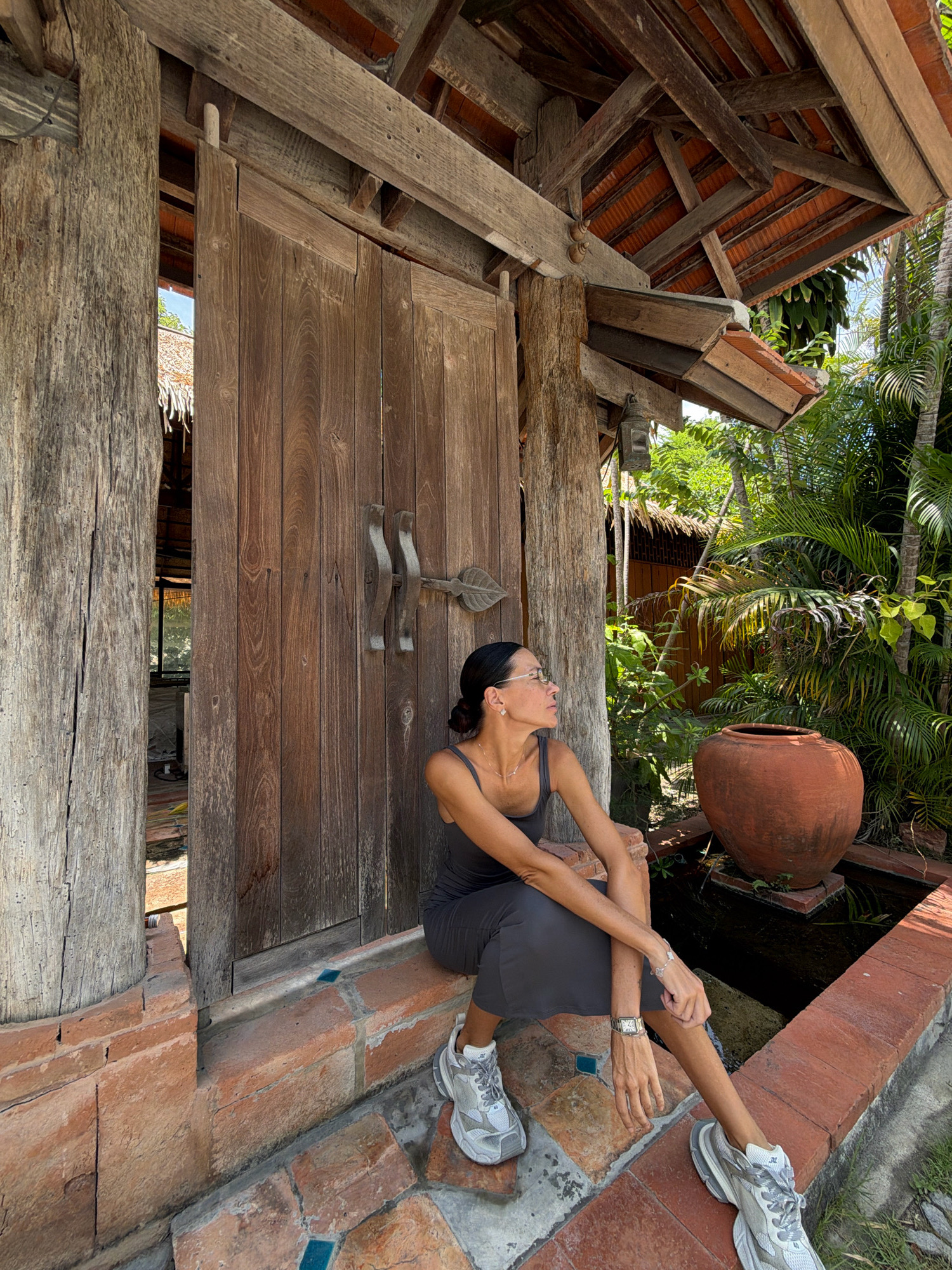 A medium shot captures a woman sitting on a brick ledge in front of a large wooden door. She is wearing a dark grey sleeveless dress and white athletic shoes. Her hair is pulled back, and she is wearing glasses and jewelry. The wooden door appears to be part of an outdoor structure, possibly a resort or temple, with exposed wooden beams and a tiled roof visible. Lush green palm trees and other tropical foliage surround the area, suggesting a warm, humid climate. A large terracotta pot sits on a water feature to the right of the woman. The ground is paved with a mosaic of stones and bricks. The scene is set outdoors in what appears to be a tranquil and natural environment, consistent with locations in Ko Samui District, Thailand.