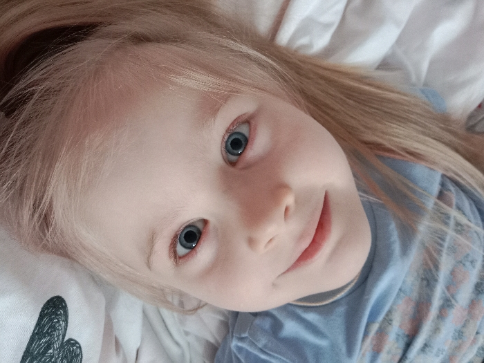 A young child with pale skin and blonde hair lies on a white surface with a gray and black heart-shaped design. The child is wearing a light blue shirt. Their bright blue eyes gaze upward, and a soft smile graces their lips. The photograph is taken from a high angle, emphasizing the child's face and expressive eyes. The lighting is soft and diffused.