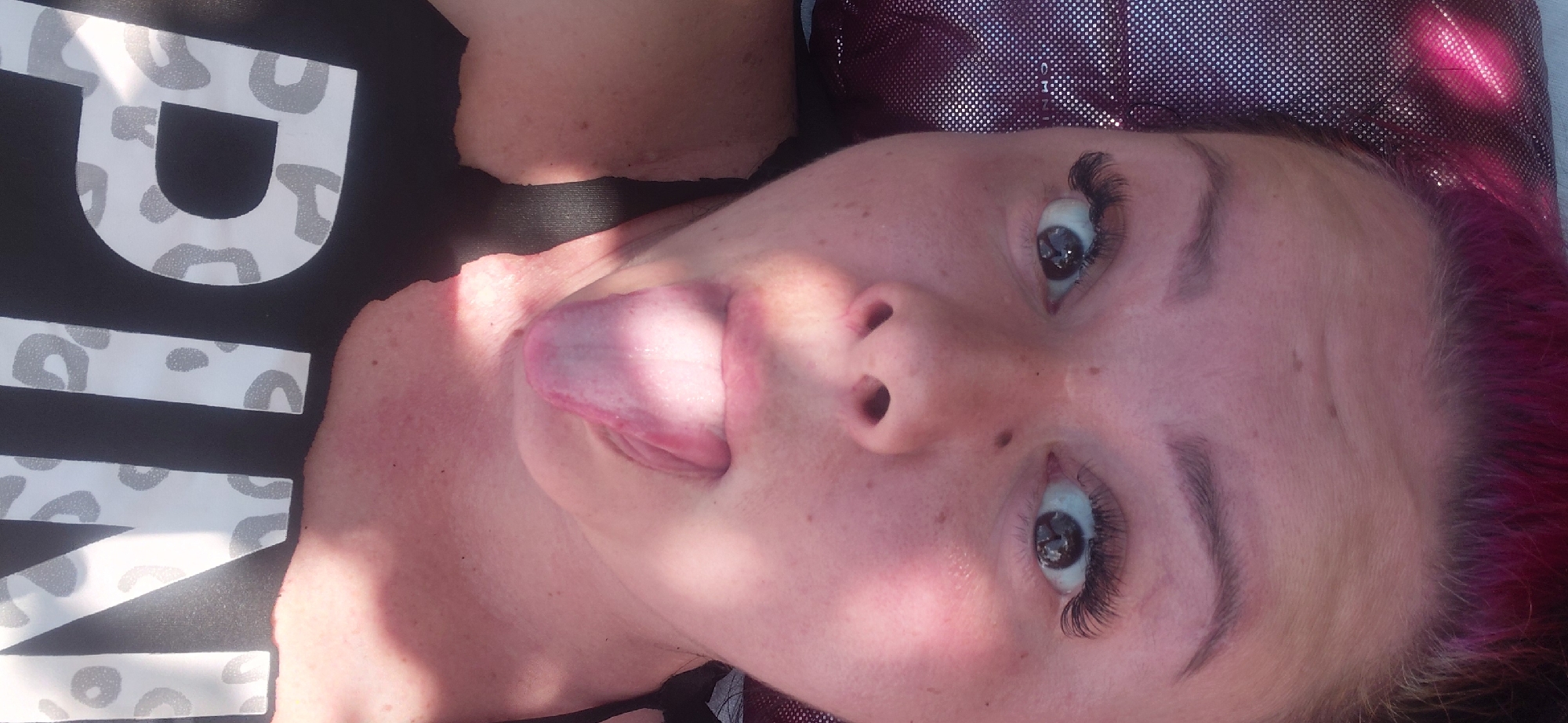 A close-up, overhead shot captures a woman’s face, her head resting on a shimmering, purple, textured surface. Her pink hair is visible at the top of the frame. She is wearing a black tank top with a white "PINK" logo. Her eyes are wide open, looking directly at the camera, and her tongue is sticking out. The scene is lit by sunlight, casting shadows across her face and neck. The location is Marynivka, Ukraine.