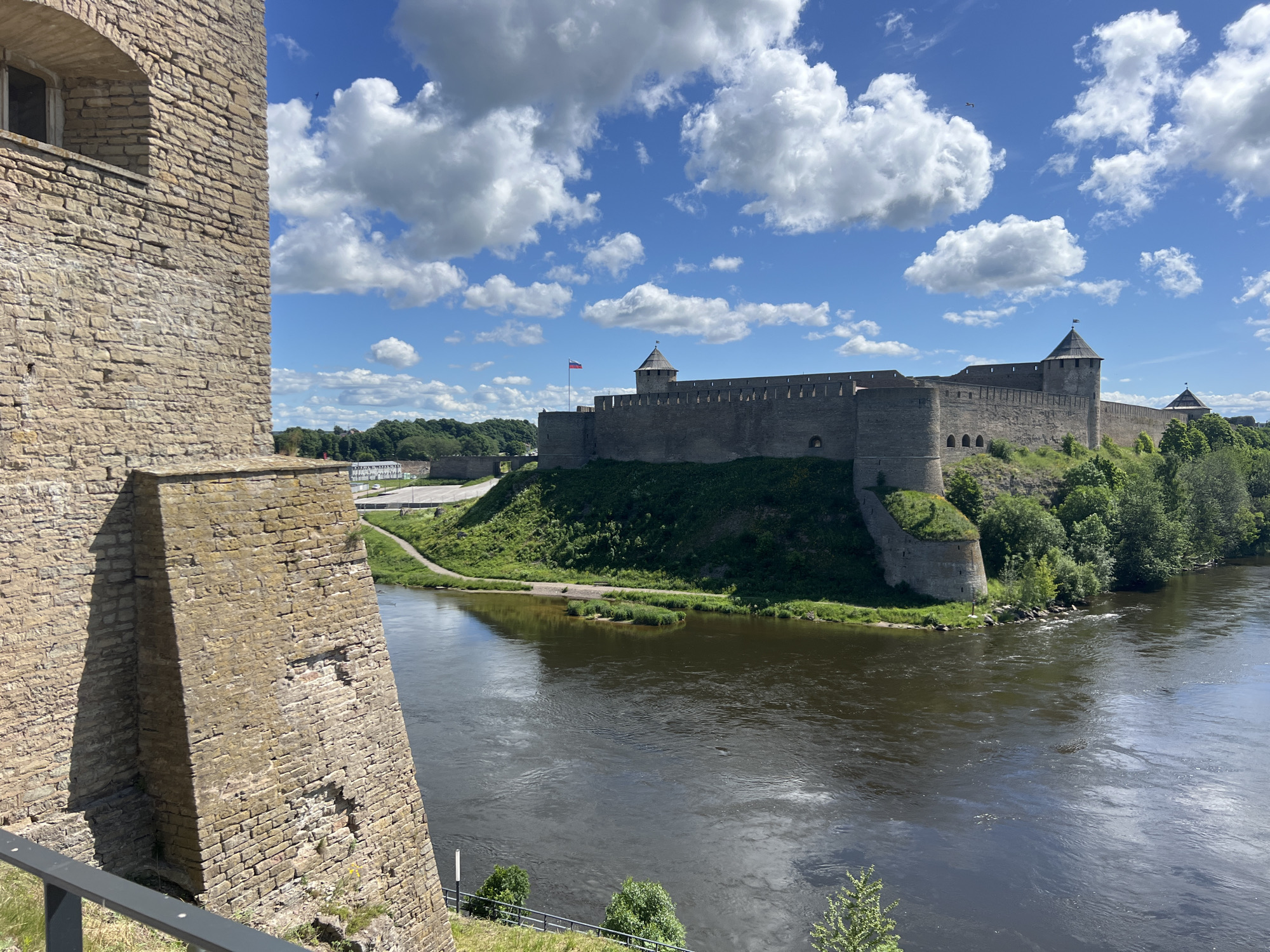 The image shows a view of Ivangorod Fortress, located on the opposite side of the Narva River in Ivangorod, Russia, from the Narva Castle (Hermann Castle) in Narva, Estonia. The foreground features a section of the stone wall of Narva Castle, with an arched window opening. The Narva River flows between the two fortresses. Ivangorod Fortress is depicted on a hill to the right of the image, with its prominent stone walls and towers. Lush green trees surround the fortress. The sky is blue with fluffy white clouds, and a flag is flying from one of the towers of Ivangorod Fortress. There are no people visible in the image.
