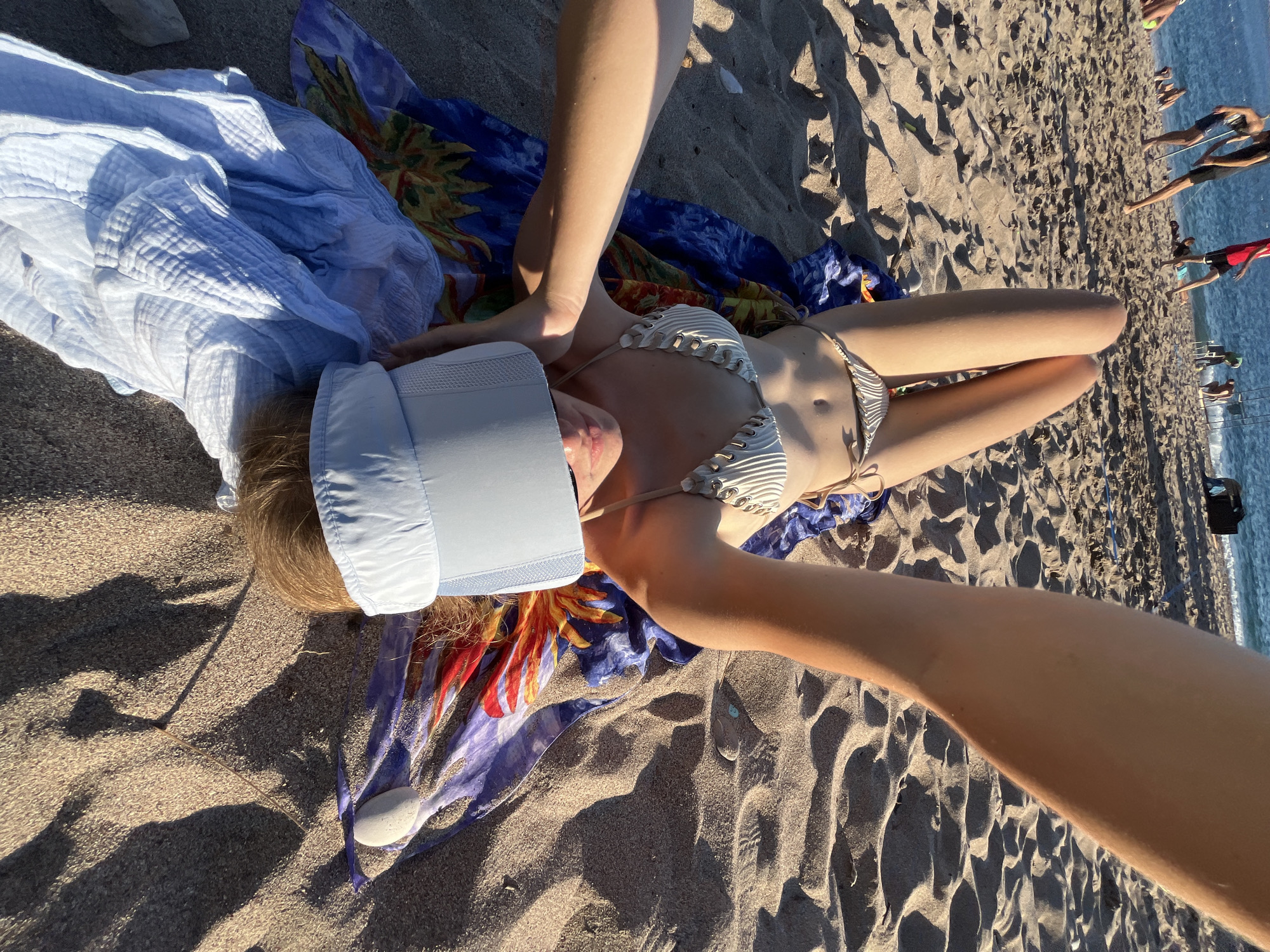 The image depicts a person lying on a sandy beach in Zankovići, Montenegro. The person is wearing a white bikini and a light blue visor. They are lying on a colorful sarong and a light blue towel. In the background, the sea is visible, with several other people in and near the water. The sun is casting shadows on the sand, indicating it is likely late afternoon or early morning.