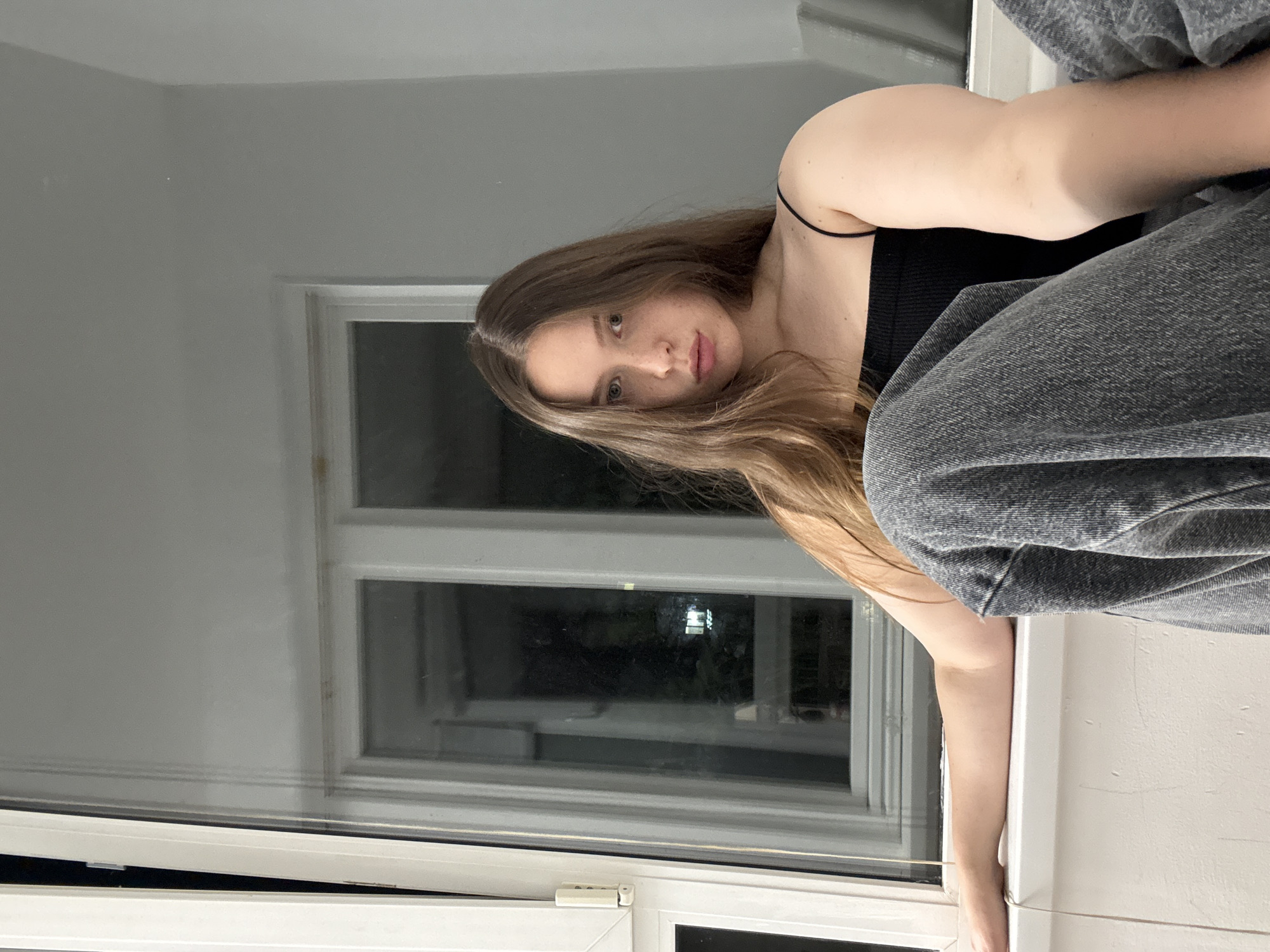 The image features a young woman with long, wavy, light brown hair, looking directly at the viewer. She has light-colored eyes and a neutral, slightly contemplative expression. She is wearing a black spaghetti strap top and grey denim pants. The woman appears to be seated or leaning on a white windowsill or ledge, with her left arm extended along its surface.
The setting is indoors, with plain light-colored walls. A large window occupies the background to her left. Through the window, the outside is dark, indicating that it is evening or nighttime, with faint artificial lights visible in the distance. The window has a white frame, and its bottom section appears slightly ajar, revealing a narrow gap. A white latch mechanism is visible on the lower part of the window frame. There is no visible text in the image. The scene is situated in Chaikovsky, Russia.