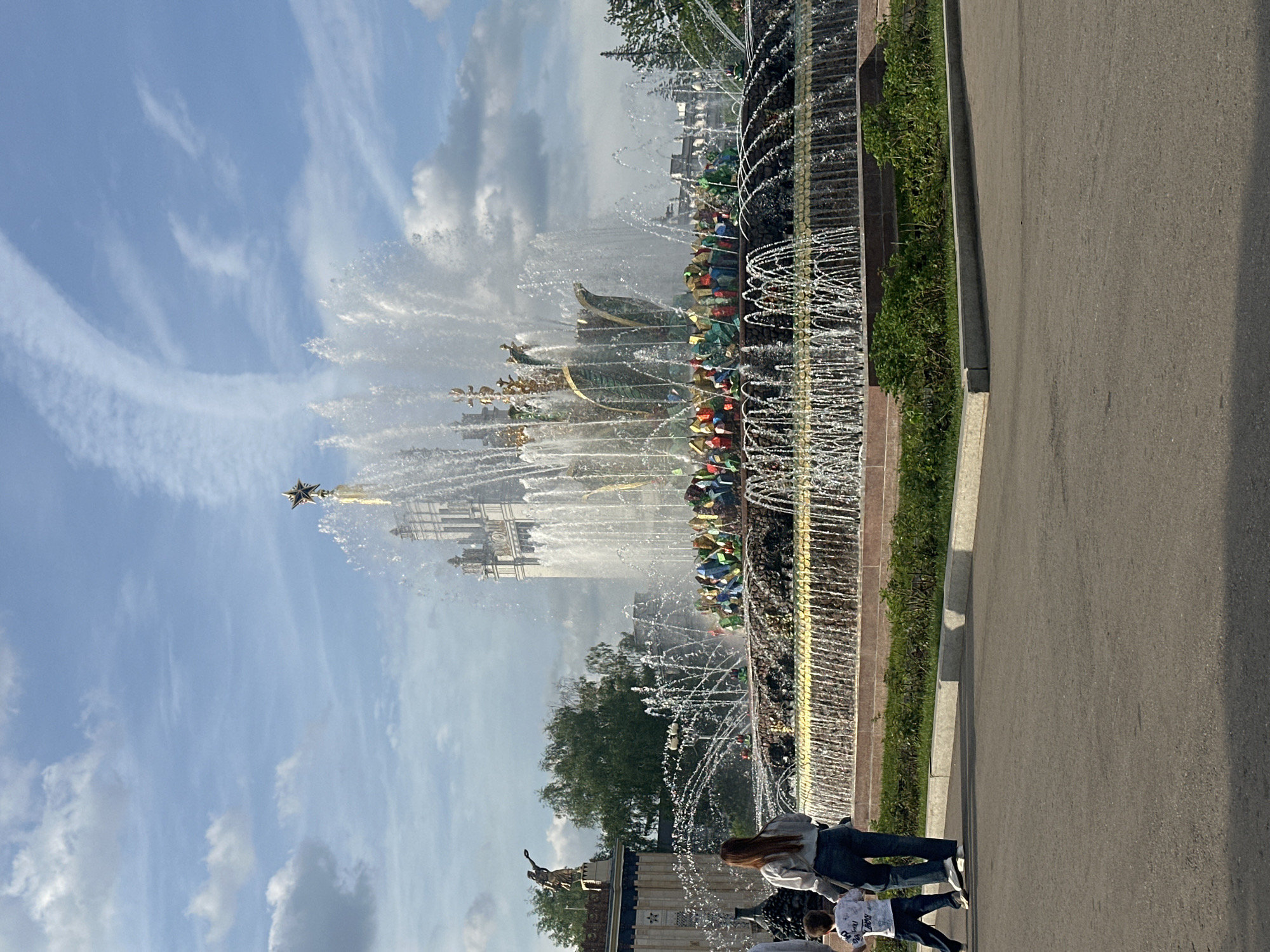 The image depicts the Friendship of Nations Fountain at the All-Russia Exhibition Center in Moscow, Russia. The fountain is adorned with golden sculptures representing different Soviet republics. Jets of water rise high into the air, creating a visually stunning display. A paved walkway is adjacent to the fountain, and people are seen walking along it. The sky is partly cloudy. Trees are visible in the background.
