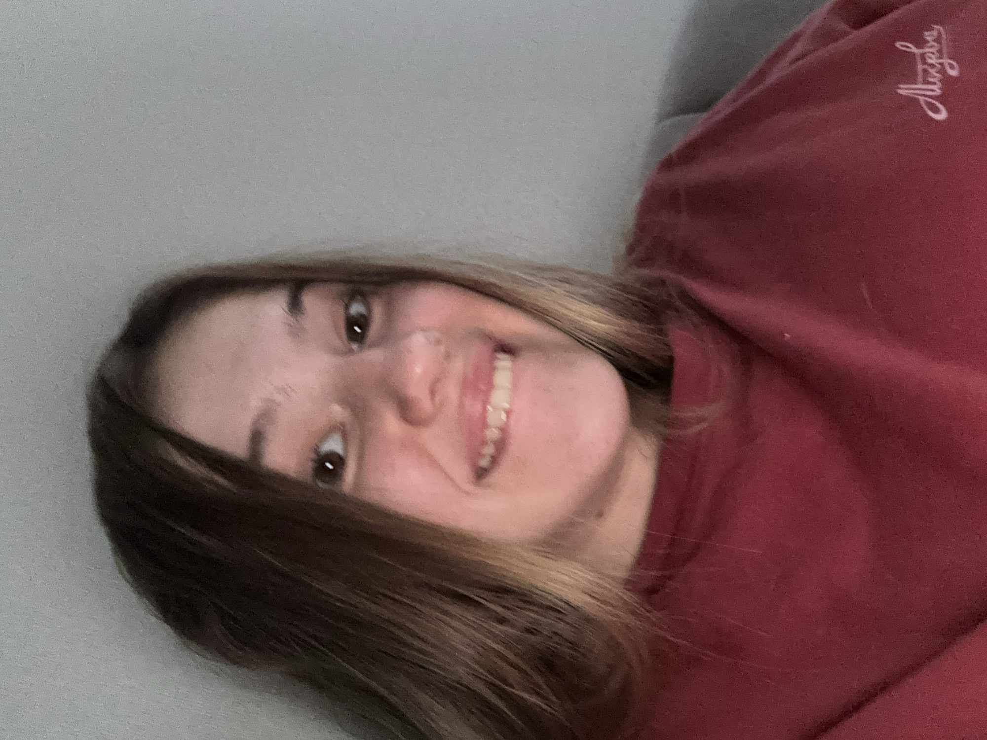 A young woman with long, brown hair smiles into the camera. She is wearing a maroon sweatshirt with a script logo on the right side of the chest. The background is a plain, light gray wall. The image is a selfie taken indoors. The location is Vsevolozhsk, Russia.