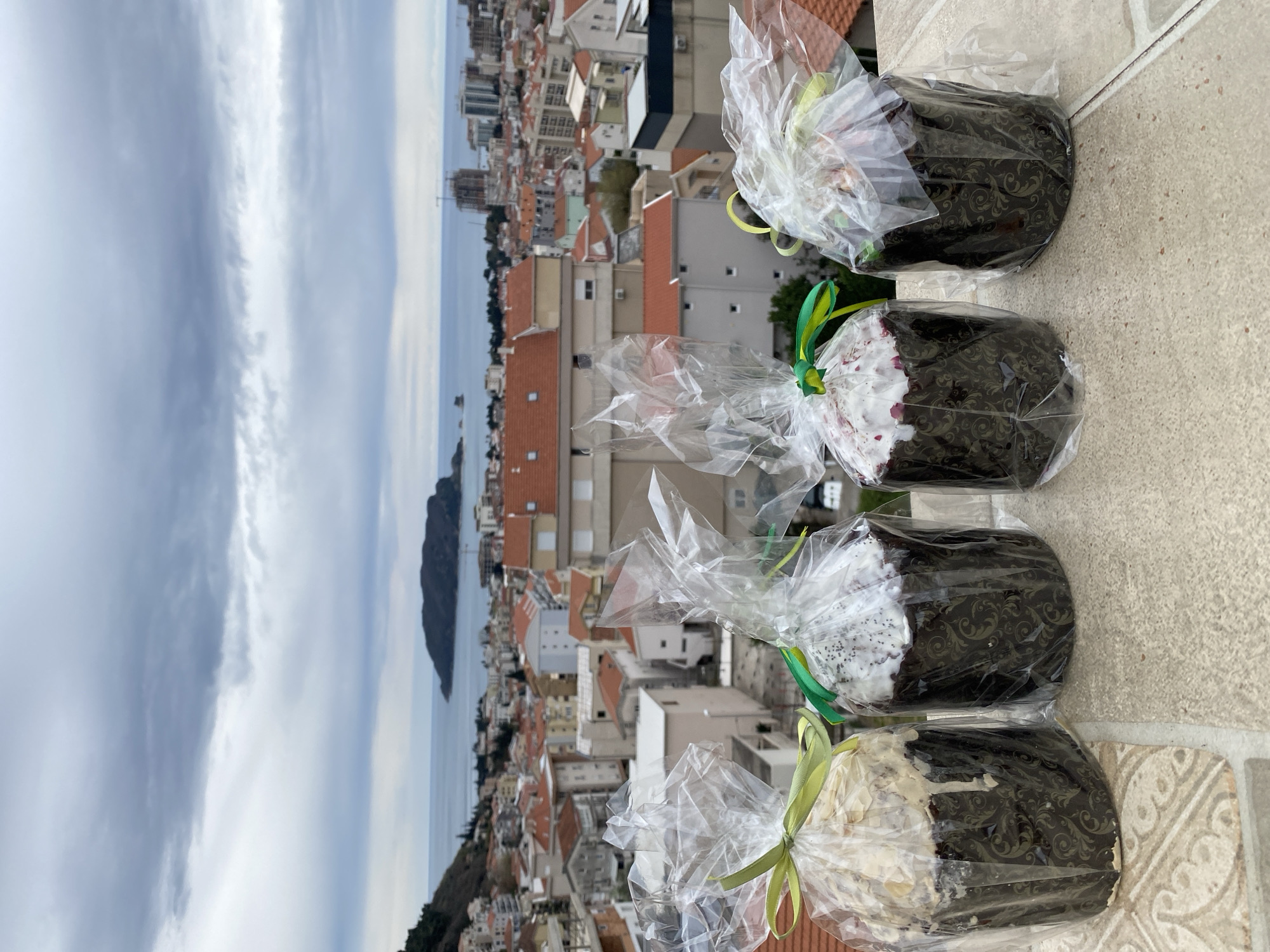 The scene is a high-angle shot, potentially taken from a balcony or a rooftop, overlooking the city of Budva, Montenegro. In the foreground, arranged on what appears to be a tiled surface, are four wrapped food items, likely pastries or cakes, decorated with white icing and tied with light green ribbons. The packaging is transparent, revealing a dark base with an intricate pattern.
The background showcases the urban landscape of Budva. Buildings with red-tiled roofs and various architectural styles densely populate the scene. In the distance, the Adriatic Sea stretches towards the horizon, with a small island visible in the water. The sky is overcast with grey clouds. The overall environment suggests a cloudy day in a coastal city. No people are visible in the frame.