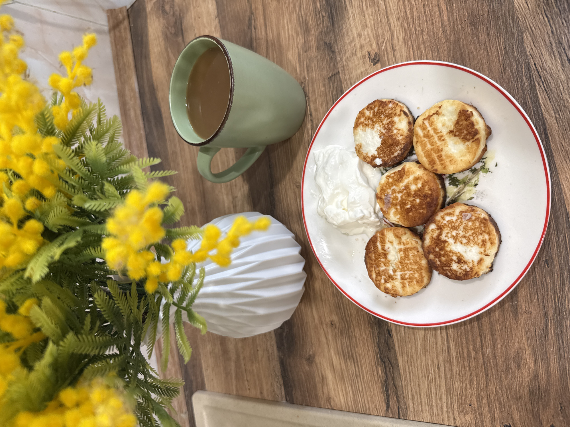A top-down shot shows a wooden table with a white plate, green mug, and a vase of yellow flowers. The plate contains five round, browned pancakes and a dollop of white cream. The green mug is filled with a light brown liquid. The vase with textured sides holds yellow flowers with green leaves. The table has a wood grain pattern. The scene appears to be indoors. The location of Rusanovo, Russia, cannot be confirmed from this image.