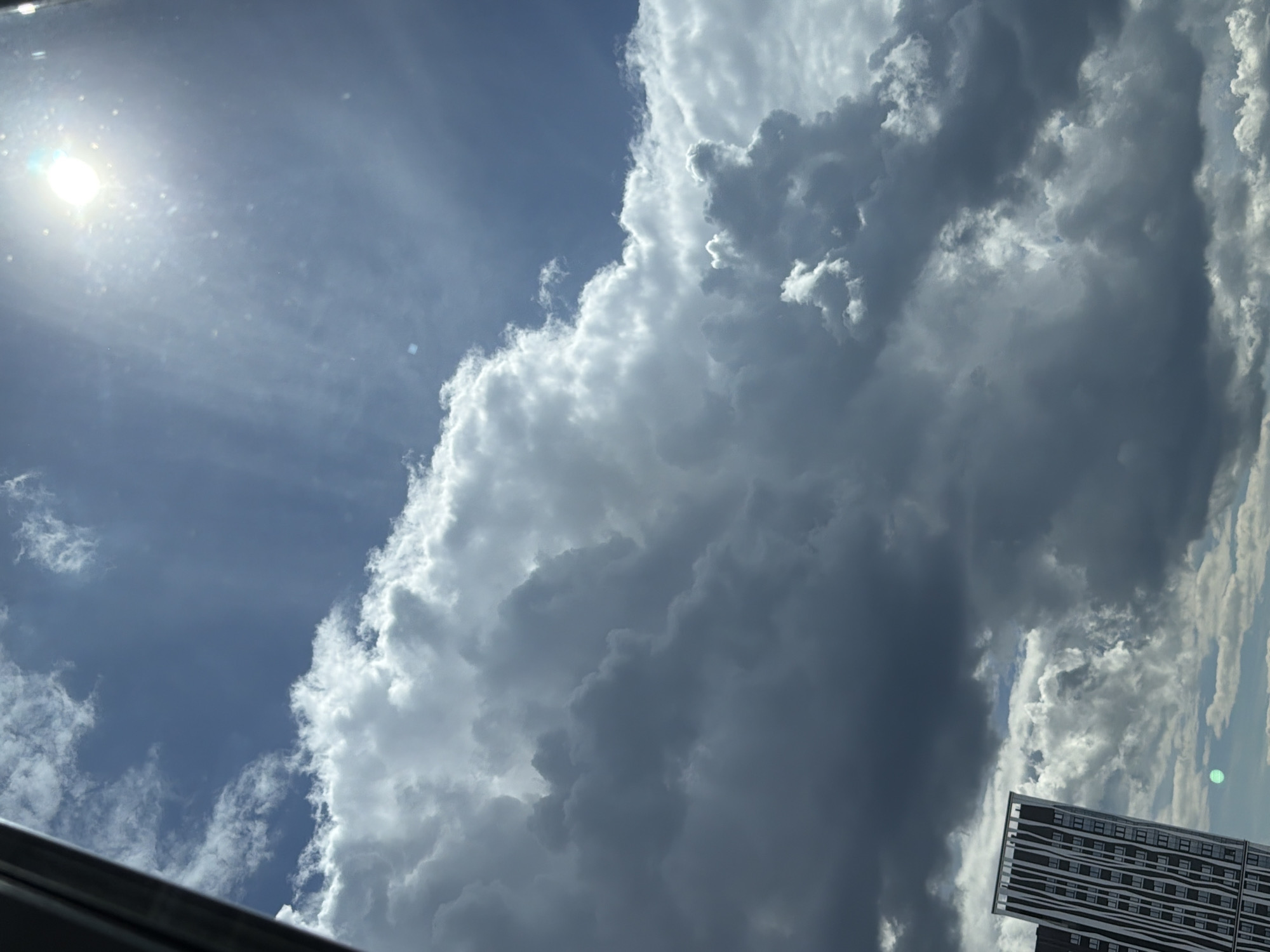 The image depicts a dramatic sky with a mix of dark, heavy storm clouds and bright, sunlit cumulus clouds. The sun is visible in the upper right quadrant, casting bright rays through the thinner clouds. In the lower left corner, the distinctive facade of a modern skyscraper is visible, characterized by vertical dark stripes on a lighter background. The building appears to be located in the city of Perm, Russia, based on the context provided. The overall scene suggests a dynamic weather event unfolding.