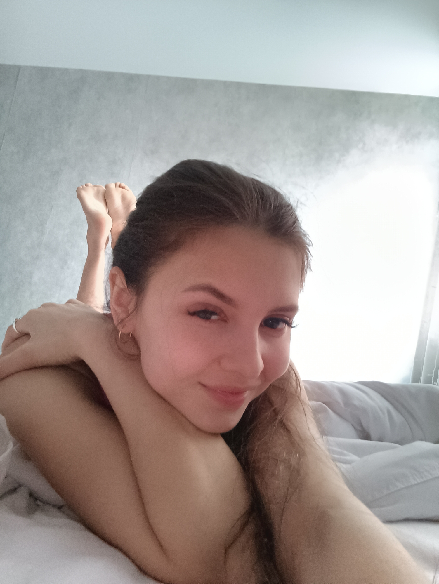 A young woman lies on a bed with white sheets. She is resting on her stomach, her arms crossed. She is smiling and looking at the camera. Her feet are raised behind her. The background is a gray wall. This appears to be an interior setting. It is not possible to confirm the location or city based on the provided image.