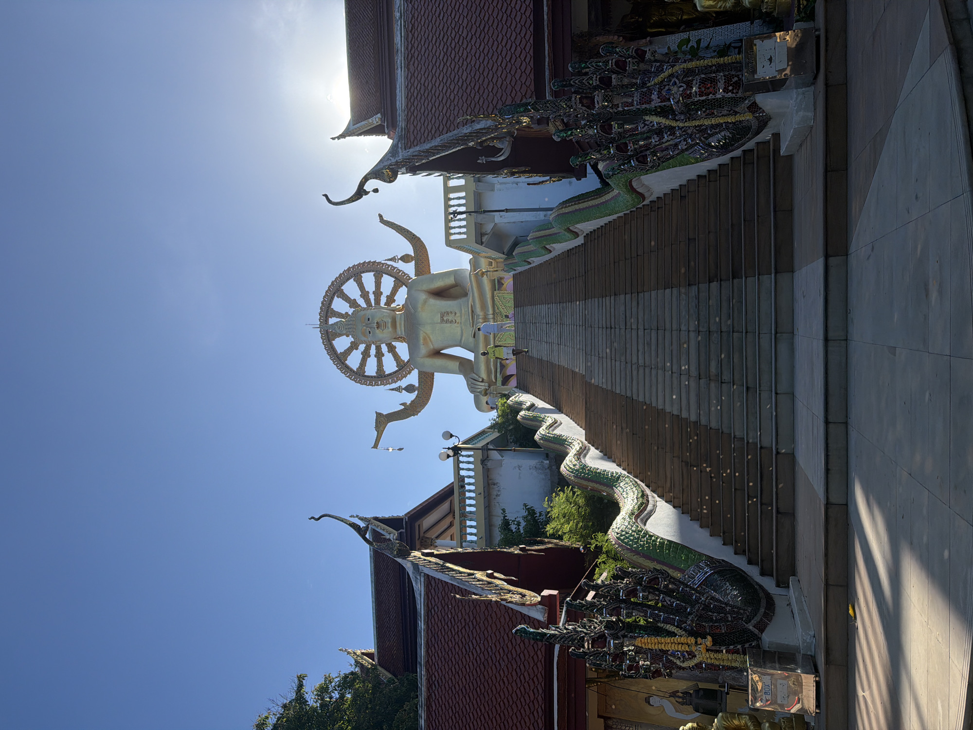 The image depicts a grand staircase leading up to a large golden Buddha statue, situated in Baan Plai Laem, Thailand. The Buddha is seated and has a Dharma wheel behind its head and multiple arms extending outwards. The staircase is flanked by ornate serpent-like railings with intricate mosaic work. Two temples with traditional tiled roofs are visible on either side of the staircase, framing the ascent. The sky is clear and blue, with the sun's glare partially obscuring the top right portion of the image. Two individuals are ascending the stairs in the distance, and the overall scene is bathed in bright sunlight, casting distinct shadows on the steps.