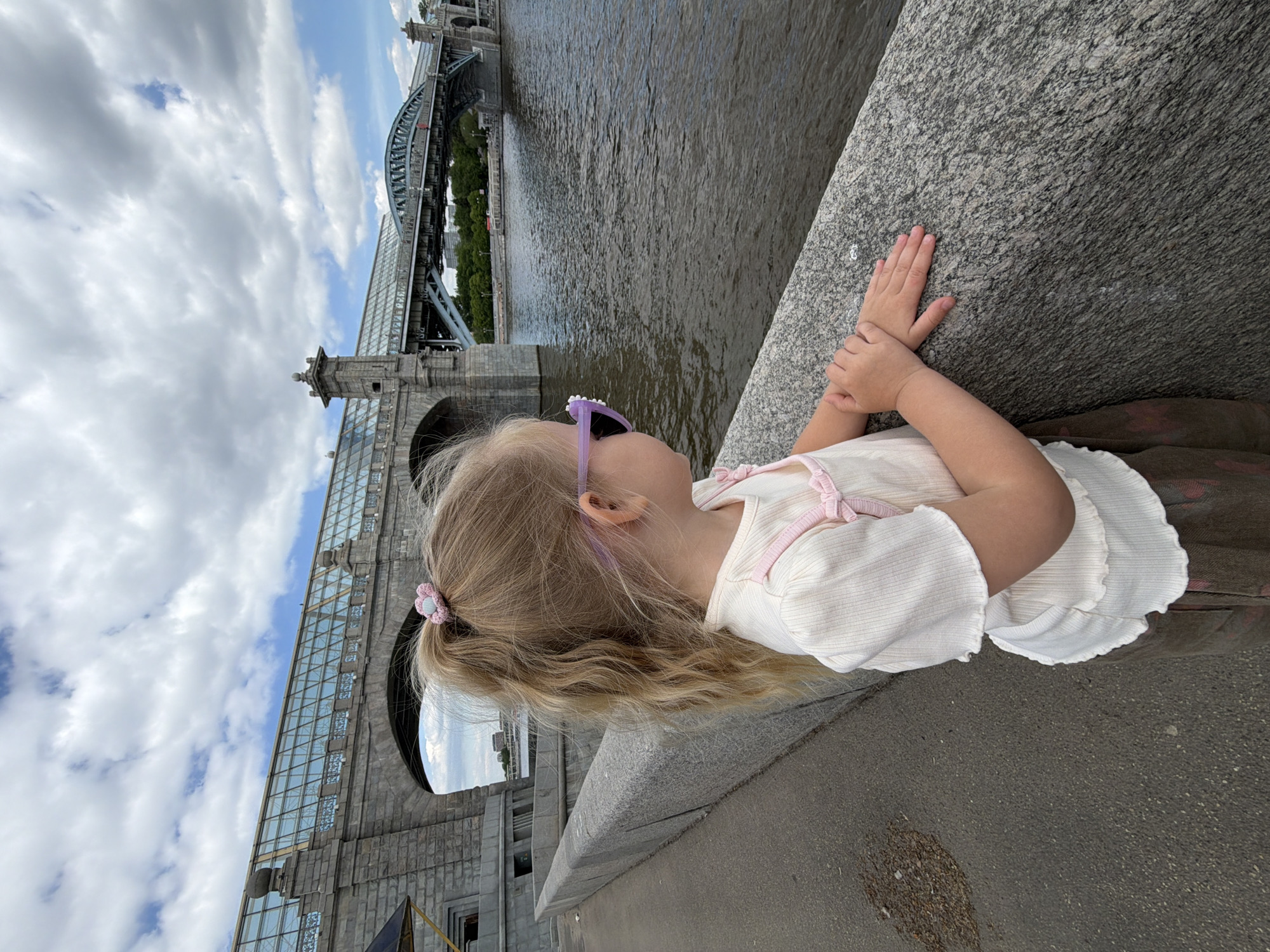 A young girl with blonde hair, wearing purple sunglasses and a white top, stands on a gray stone ledge. Her hands rest on the ledge as she looks out towards a river. In the background, there's a complex bridge structure made of gray stone and steel, with multiple arches and towers. The sky is blue with scattered white clouds. The scene appears to be in Moscow, Russia, given the architectural style of the bridge, which resembles the Luzhniki Metro Bridge. The environment includes both the urban architecture and the natural element of the river.