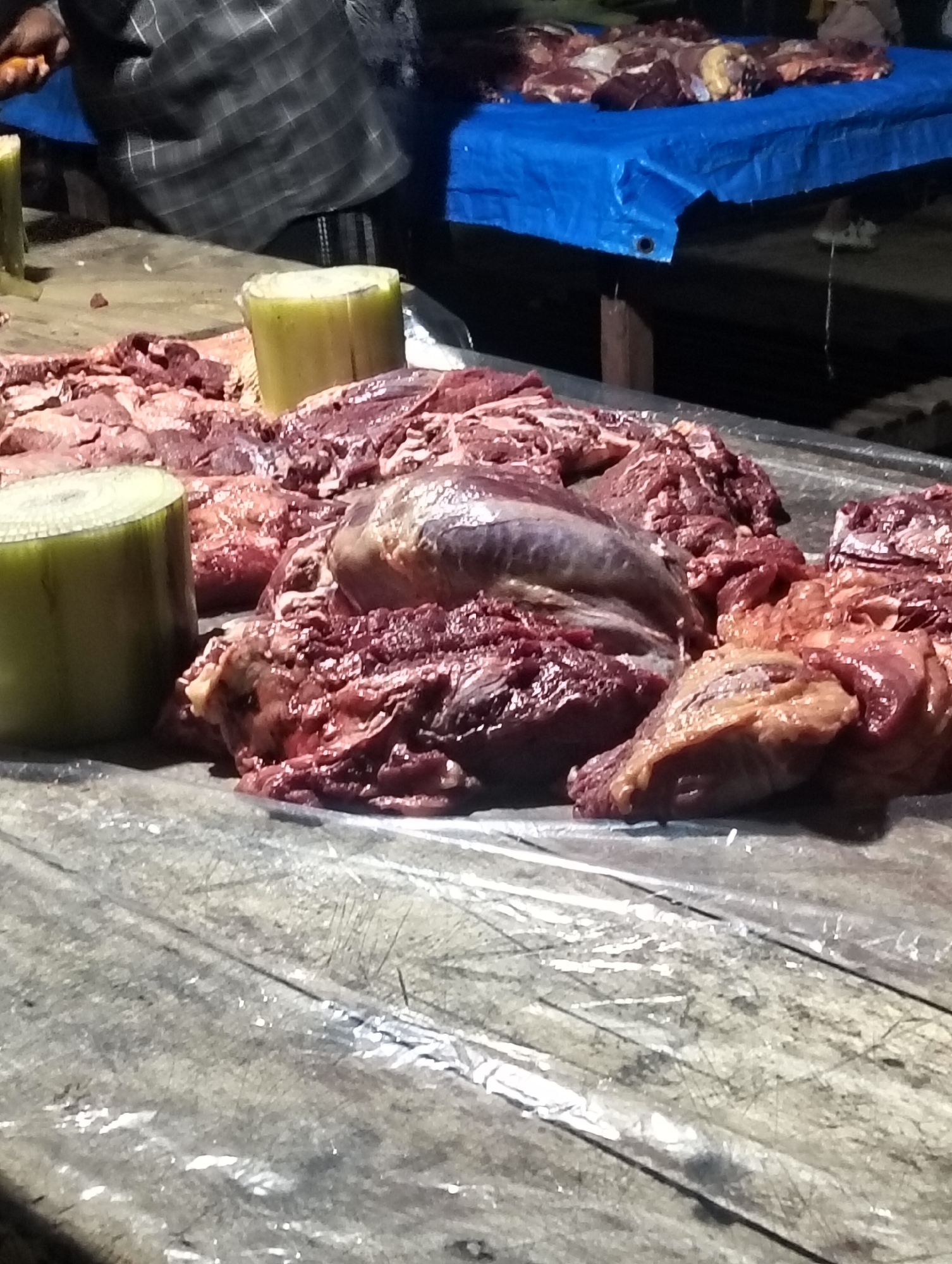 This media file depicts a scene at a market in Cottrueng, Indonesia, likely a butcher's stall. The primary subjects are large cuts of raw, red meat, piled on a wooden table covered with a plastic sheet. Two cylindrical, pale green objects, possibly stalks of banana tree, are placed near the meat. In the background, a person wearing a checkered shirt is partially visible, with their hands appearing to be engaged with the meat or preparing the stall. A blue tarp is spread over a surface in the background, suggesting a covered market area or vendor's setup. The lighting suggests it could be daytime or a well-lit indoor space. The overall impression is one of a busy, active marketplace where raw meat is being sold. There is no visible text in the image.