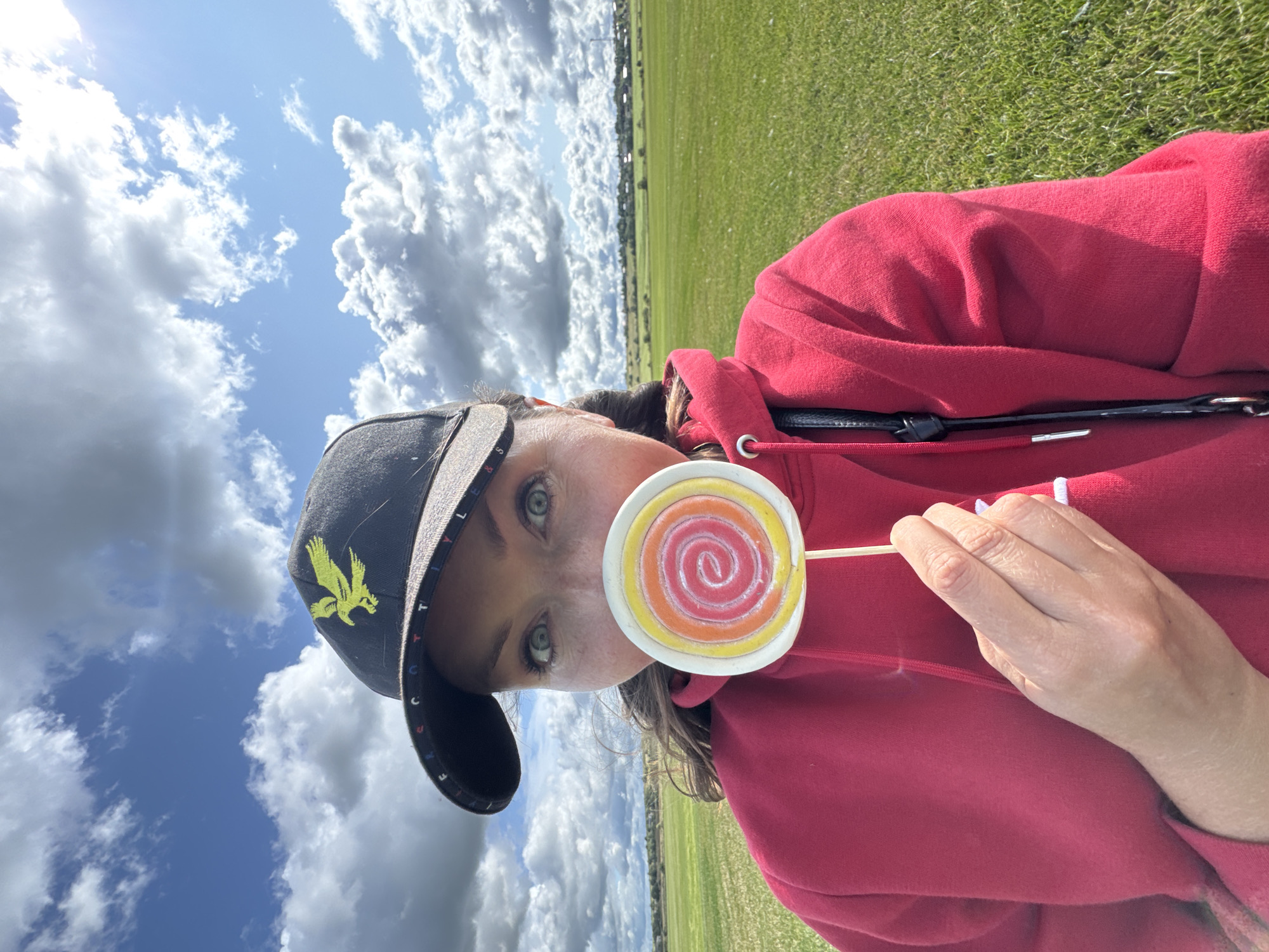 A person is taking a selfie outdoors in a grassy field under a cloudy sky. The person is wearing a red hooded sweatshirt and a black baseball cap with a yellow bird logo. They are holding a colorful swirl lollipop in front of their mouth, obscuring it from view. The background shows a flat green landscape with a treeline and buildings in the distance. The overall lighting suggests it is daytime.