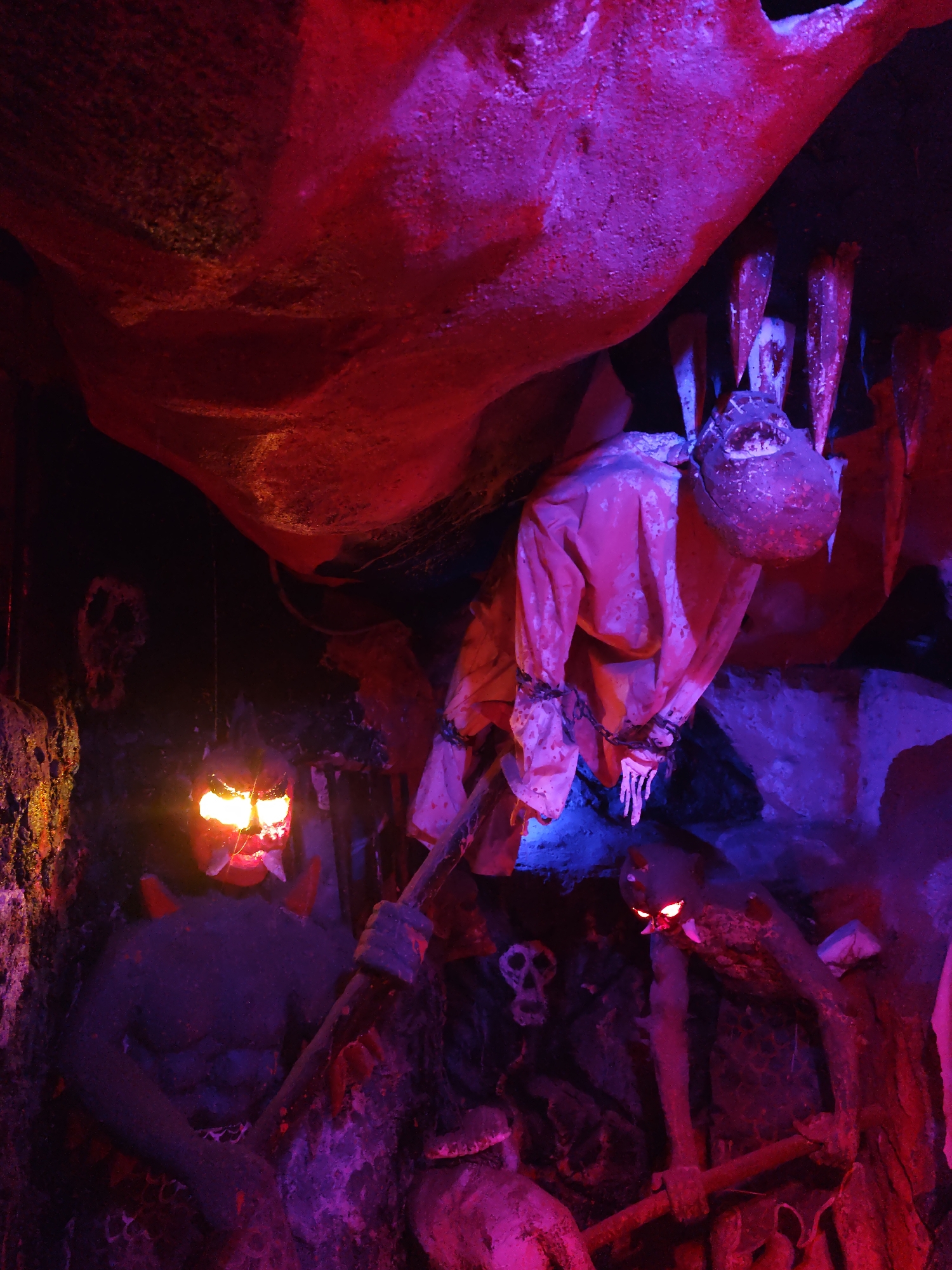 The image depicts a dark, cavernous environment with theatrical lighting. In the foreground, a large, dark figure with glowing red eyes and two horns is visible. This figure appears to be holding a wooden pole. To the left of this figure, another dark figure with glowing yellow eyes and prominent red horns is positioned. This figure also holds a wooden pole and has a menacing expression. In the midground, a macabre scene unfolds with a suspended, shrouded figure in a pink garment, bound by chains. Its head is indistinct, adding to the unsettling atmosphere. Scattered throughout the scene are elements suggestive of a hellish or macabre setting, including what appear to be bones or skulls and jagged, rock-like structures illuminated by vibrant red and purple lights. The overall impression is one of a staged horror exhibit or a scene from a dark fantasy. The location is unknown, and there are no discernible elements that place it in a specific city or region.