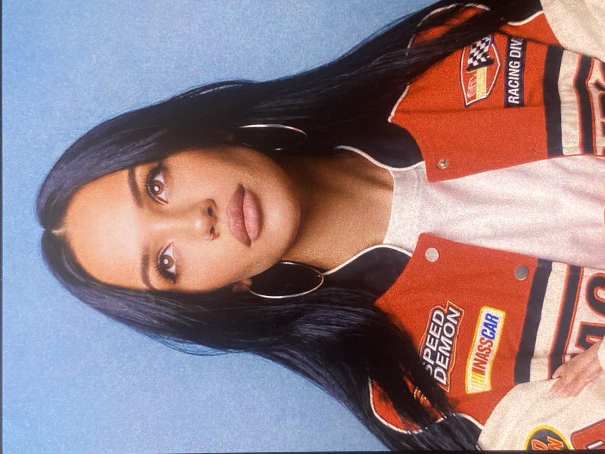 A close-up portrait of a young woman with long, dark hair and light brown eyes, wearing a red and white racing-themed jacket over a white t-shirt. She is looking upwards and to her right, with large hoop earrings visible. The background is a solid light blue. The image appears to be a studio portrait with a plain backdrop.