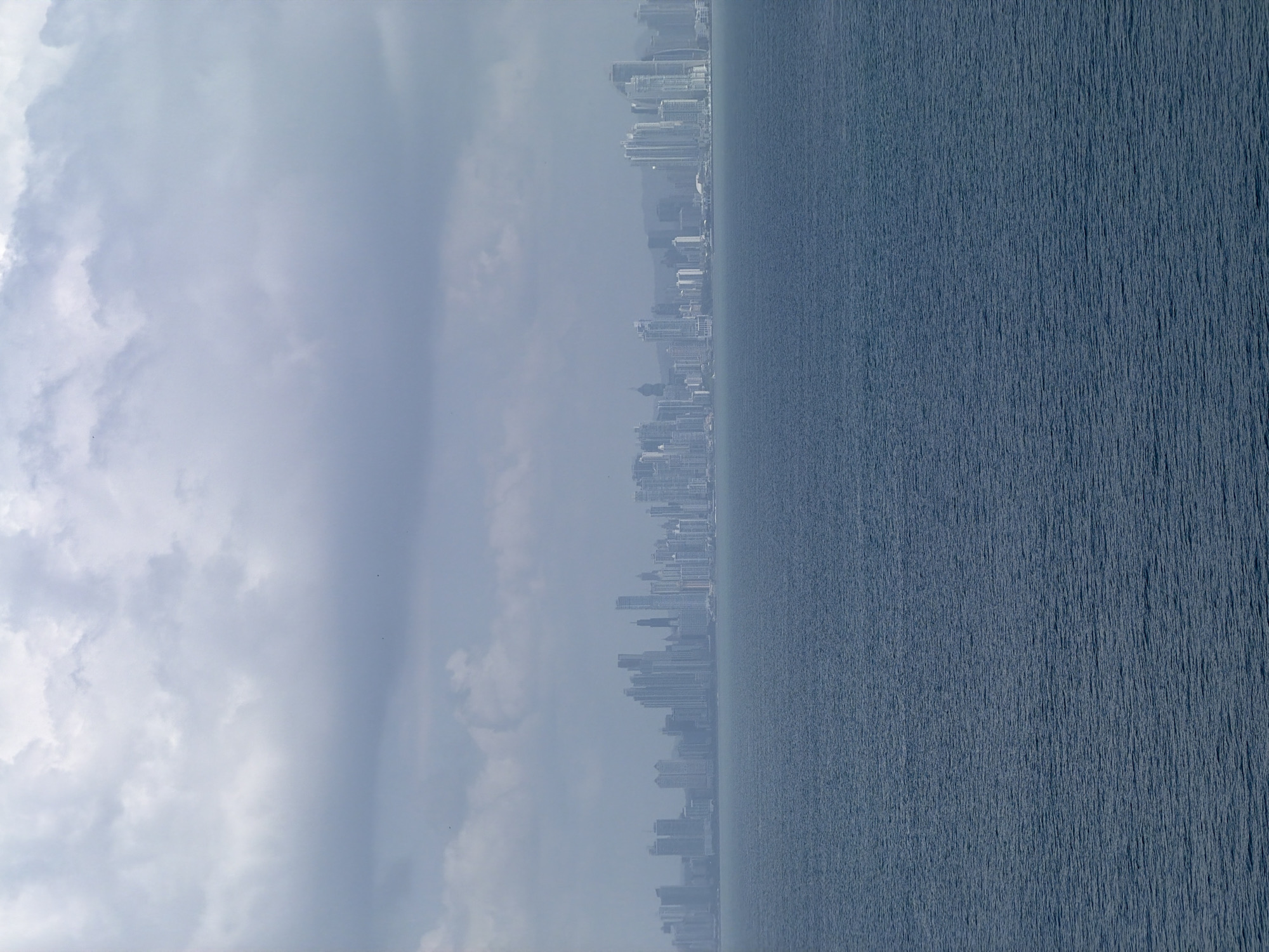 This is a wide-angle, eye-level shot of a distant cityscape on the horizon, separated from the viewer by a large body of water. The sky above is overcast with heavy, gray clouds. The water in the foreground is a dark, choppy blue. The cityscape, partially obscured by haze, features a dense collection of modern skyscrapers of varying heights and architectural styles, predominantly white or light-colored. The buildings appear densely packed, suggesting a significant urban center. Based on the characteristic skyline, this is the city of Panama City, Panama, specifically the financial district with its numerous high-rise buildings. No people are visible in the image. The overall atmosphere is subdued and slightly melancholic due to the cloudy weather.