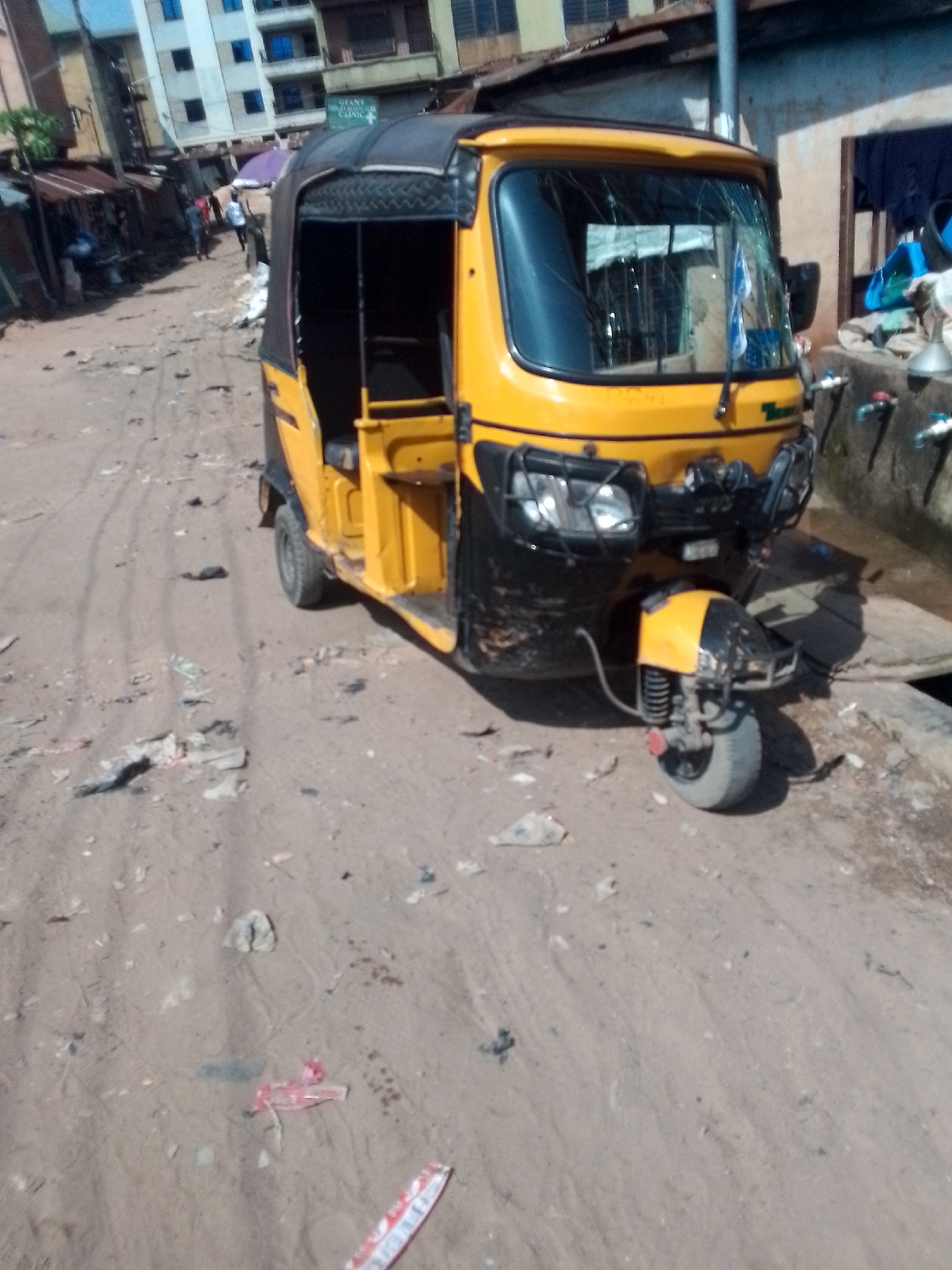 The image depicts a street scene in Onitsha, Nigeria, characterized by a dusty, unpaved road littered with debris. In the foreground, a yellow and black auto-rickshaw, commonly known as a "keke," is parked. The vehicle has a covered passenger compartment and a visible driver's seat. The front of the keke features two headlights and a protective metal bar. Its front wheel is turned to the right, and it appears to be stationary.
In the background, several multi-story buildings line the street, suggesting an urban or semi-urban setting. The architecture of the buildings is varied, with some appearing to be modern with blue-tinted windows, while others seem older and more weathered. A few individuals are visible walking on the street in the middle ground, blurred by the distance and the image resolution, indicating some pedestrian activity. Shops with corrugated metal roofs are also present along the roadside, implying commercial establishments. The overall environment suggests a lively but somewhat neglected urban area, typical of many Nigerian cities.