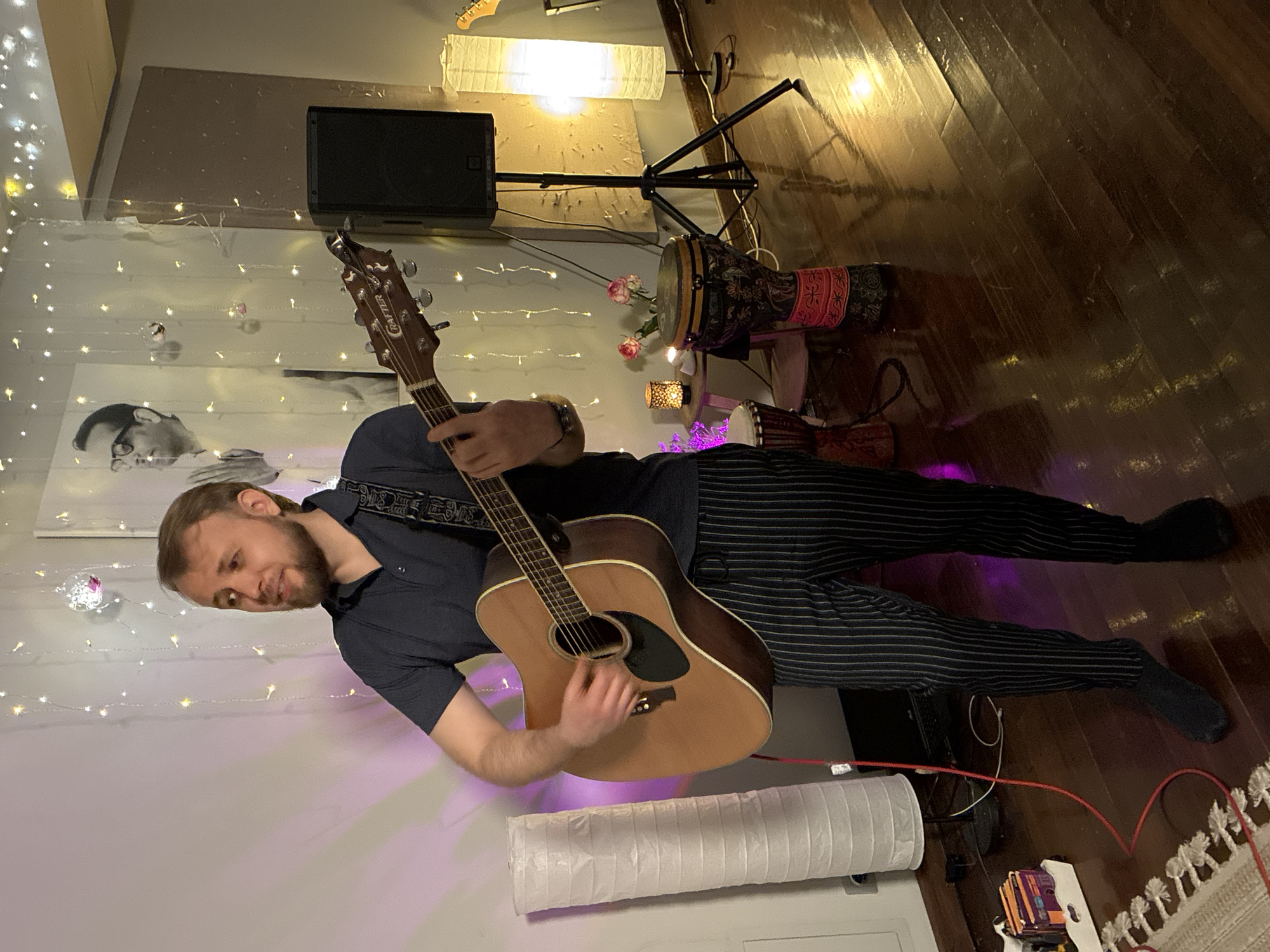 An adult male in his late 20s to early 30s, with light brown hair and a short beard, stands centrally in an indoor setting in Moscow, Russia. He wears a dark blue polo shirt, dark pants with thin white vertical stripes, and dark socks. The man is actively playing a natural wood acoustic guitar with a black pickguard and a patterned strap, his right hand strumming near the soundhole and his left hand on the fretboard. His mouth is slightly open, suggesting he is singing or speaking, and his gaze is directed upwards and to the left.
The background features a light-colored wall adorned with multiple strands of warm-toned string lights and reflective ornaments. A large black and white portrait of a man in glasses with clasped hands is visible on the wall behind the performer. To the right, a black PA speaker stands on a black tripod. A small round table positioned between two djembe drums holds two illuminated candle lanterns and a small vase containing two pink roses. The djembe drums vary in size, with the larger one featuring colorful patterns and a red fabric band. Part of another guitar's headstock and neck is visible on the far right.
To the left of the performer, a tall white paper lantern-style floor lamp stands unlit, near a white tray containing several small rectangular objects and a partially obscured black laptop on the dark wooden floor. A prominent red audio cable extends from the guitar towards the floor and to the left. Another similar white paper lantern-style floor lamp on the far right is illuminated, casting a warm glow. The polished dark hardwood floor reflects the room's lighting. A patterned rug is partially visible in the bottom-left foreground. Purple ambient lighting emanates from the lower left, casting a hue on the wall behind the performer.