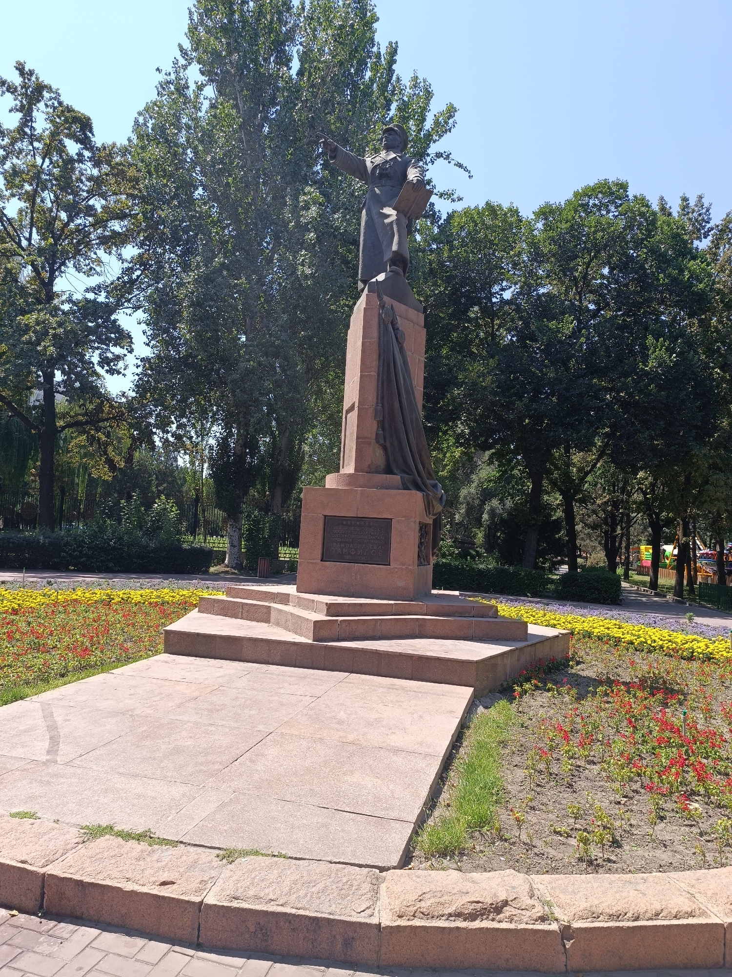 This is a full-shot, eye-level, outdoor photograph of a monument in Bishkek City, Kyrgyzstan. The monument is made of dark bronze and stone, depicting a figure standing atop a pedestal and pointing forward with one hand, while holding a book in the other. The pedestal is adorned with a draped cloth. The monument is surrounded by a lush green park setting with mature trees. In the foreground, there are two flower beds, one filled with yellow flowers and the other with red flowers. The ground around the monument is paved with stone tiles and bordered by a stone curb. The sky is clear and blue. The monument is located in Oak Park in Bishkek, Kyrgyzstan, which is a historical park with several monuments.