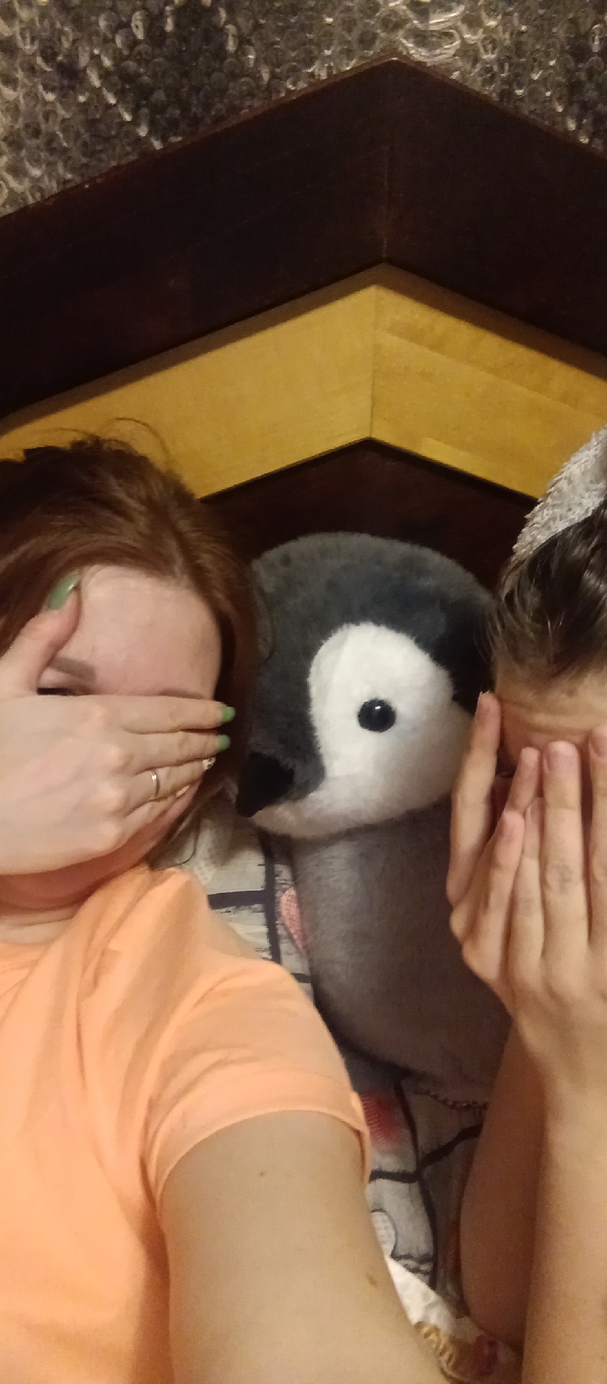 Two people and a stuffed penguin are in the scene. Both people are covering their eyes with their hands. The person on the left is wearing an orange shirt and has light green nail polish. The person on the right has a hair accessory. They are positioned in front of a wooden headboard. A patterned blanket is visible beneath them. The location is Unknown, Unknown.