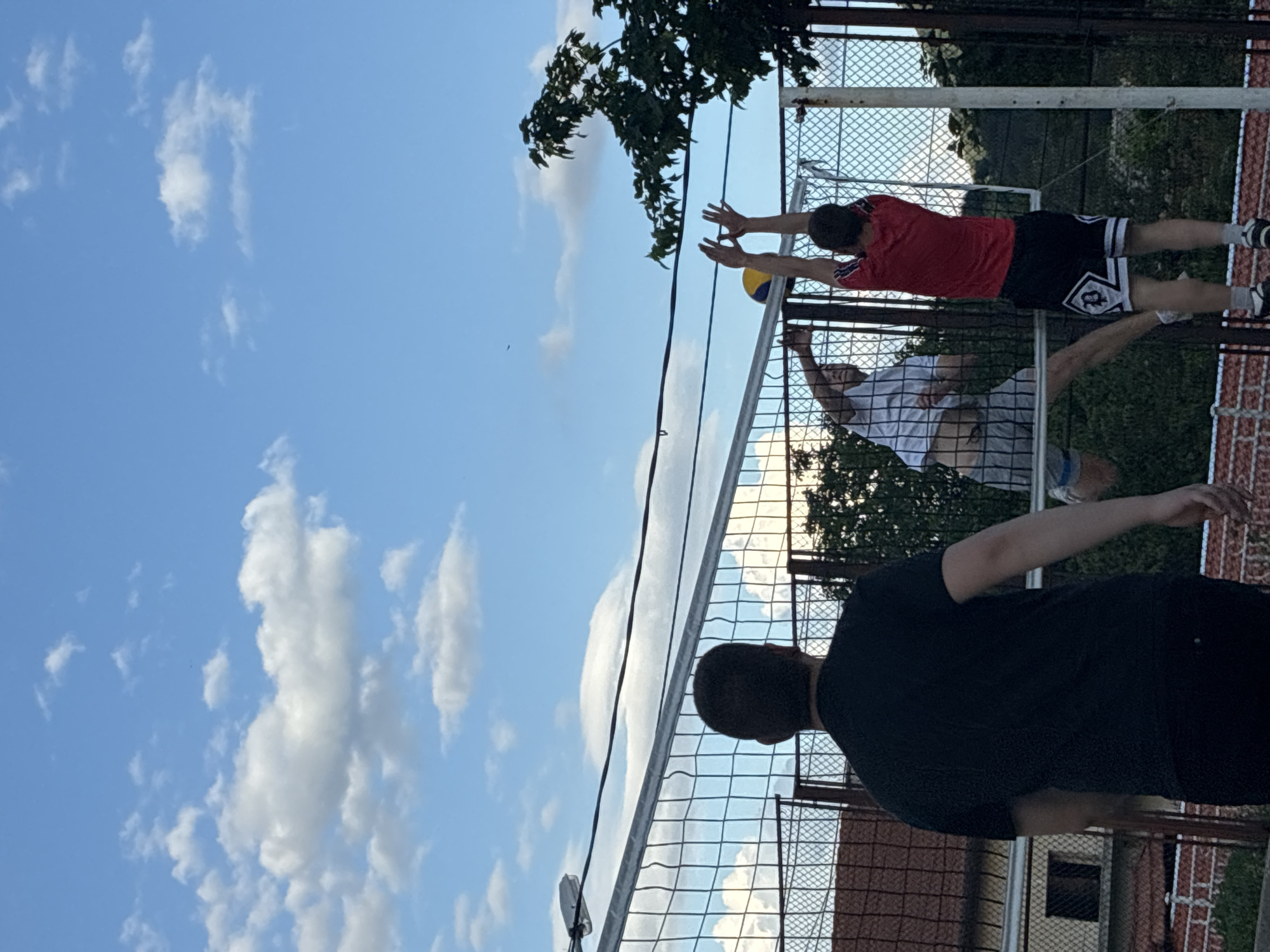 The image captures an outdoor volleyball game in Klisura, Bulgaria, under a clear blue sky with scattered white cumulus clouds. In the foreground, viewed from behind, a person in a black t-shirt and dark pants stands observing the scene. A volleyball net stretches horizontally across the midground, separating the court. On the far side of the net, two male players are airborne. The player on the left, wearing a white t-shirt and grey shorts, has his left arm extended over the net. To his right, another player in a red t-shirt and black and white shorts is mid-jump, with both arms raised above his head, hands positioned to play the yellow and blue volleyball visible directly above the net. Behind the players and net, a chain-link fence is visible, with a building featuring a red tiled roof and a dark window partially seen on the left. On the right, green trees and foliage are present. Overhead power lines and a street light fixture are visible in the upper portion of the frame.