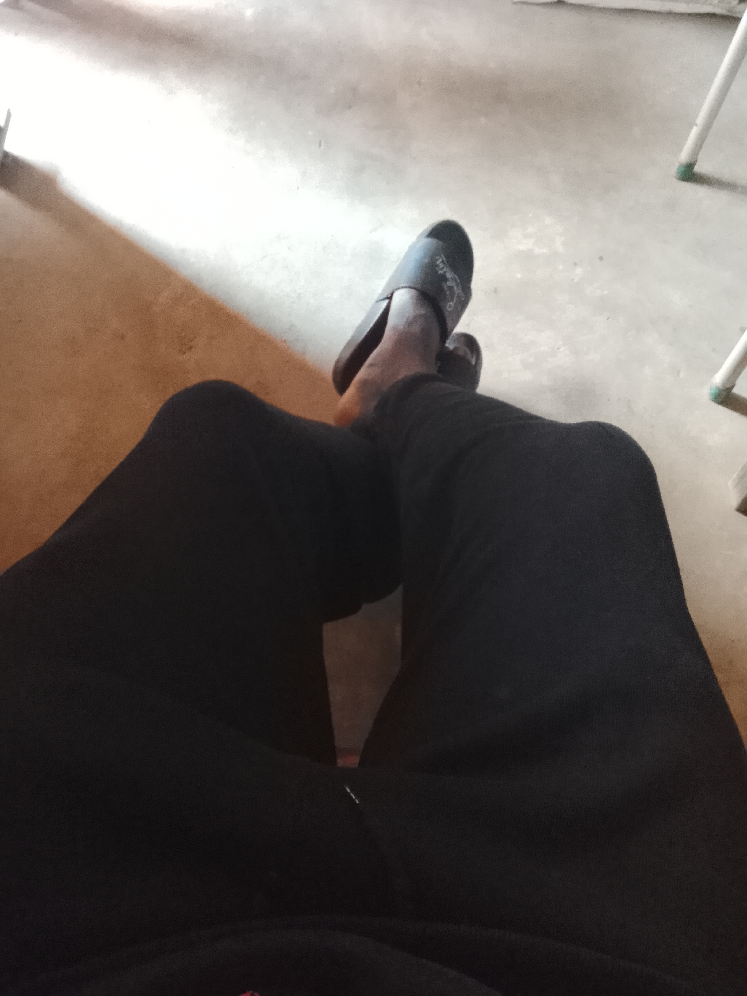 The image shows a person's legs and feet, wearing dark pants and dark slide sandals. The person is sitting on a floor that appears to be concrete or a similar hard surface, with a lighter colored, possibly wooden, surface on the left. The background is blurred and shows parts of what appear to be chair legs. The scene suggests an indoor or covered outdoor setting. There is no specific indication of a location within the city of Durumi, Nigeria.