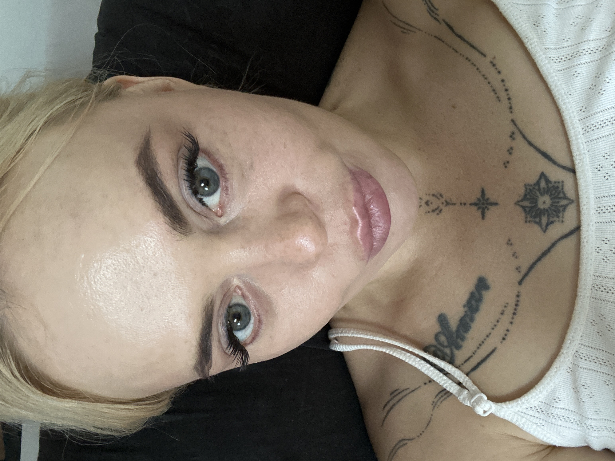 An overhead, close-up, forensic shot shows a fair-skinned woman with blonde hair, light blue eyes, and full lips, wearing a white tank top. She is positioned with her head tilted slightly to her right, looking directly into the camera. Her skin is clear with light freckling and has a natural sheen. Her eyebrows are dark and well-defined, and her eyelashes are long and voluminous, with noticeable mascara. She is wearing light pink lipstick. Her left shoulder and upper chest are visible, adorned with several black tattoos. One tattoo on her upper chest reads "Christian" in cursive. Another tattoo, centered below it, is a stylized star or floral pattern. Delicate line tattoos extend upwards from her collarbone towards her neck. The background is a dark, textured surface, likely a sofa or cushion, which is out of focus. The lighting is even, highlighting the woman's features and the details of the tattoos. The image is sharp and well-exposed, providing clear detail of the subject's face and upper body. No specific location in a city is identifiable from the image.