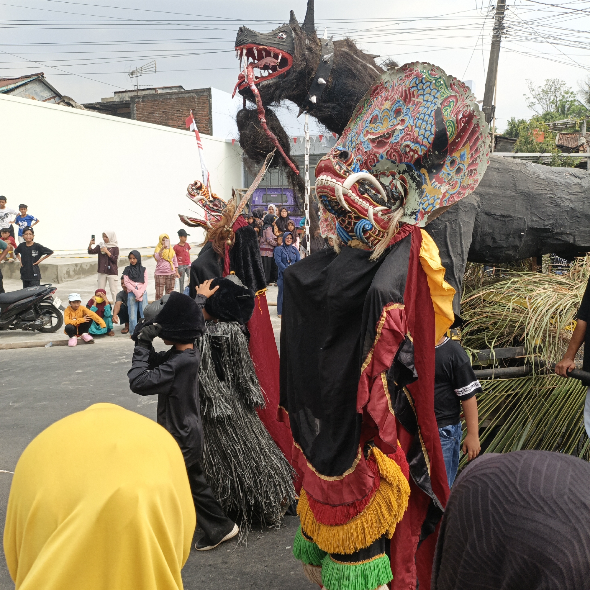 This is a medium-shot, eye-level photograph taken outdoors in Randudongkal, Indonesia, during what appears to be a festival or cultural event. The scene is dominated by two large, elaborately decorated figures. The one on the right is a masked humanoid figure wearing a black robe with red and gold trim, featuring layers of red, yellow, and green fringe at the hem. A large, colorful, dragon-like mask adorns its head. To the left is another figure in a black robe and a black, furry headpiece, holding a smaller, similar mask. Behind these figures, a massive, dark, dragon-like effigy with a gaping mouth and red teeth is visible, along with another dragon's head to its left, adorned with a red and white striped flag.
A crowd of onlookers, including many children and some adults, are gathered in the background, some holding up their phones to take pictures. Several buildings, including a white wall and some structures with corrugated metal roofs, are visible behind the figures and crowd. A black motorcycle is parked to the left of the frame. The ground is paved, and there are overhead power lines and a utility pole in the upper portion of the image. The overall atmosphere suggests a lively public celebration.