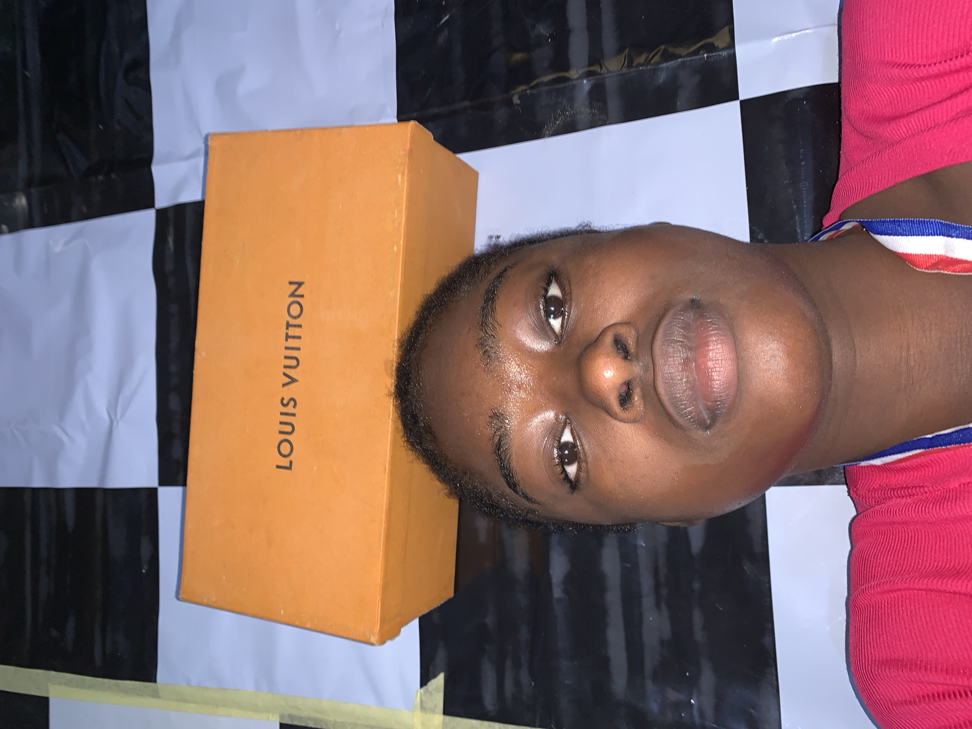 A person lies on a surface patterned with black and white squares, wearing a pink top with a striped blue and red trim. Next to the person's head is an orange box with "LOUIS VUITTON" printed vertically on its side. The location is inferred to be in the city of Rafin Par, Nigeria.