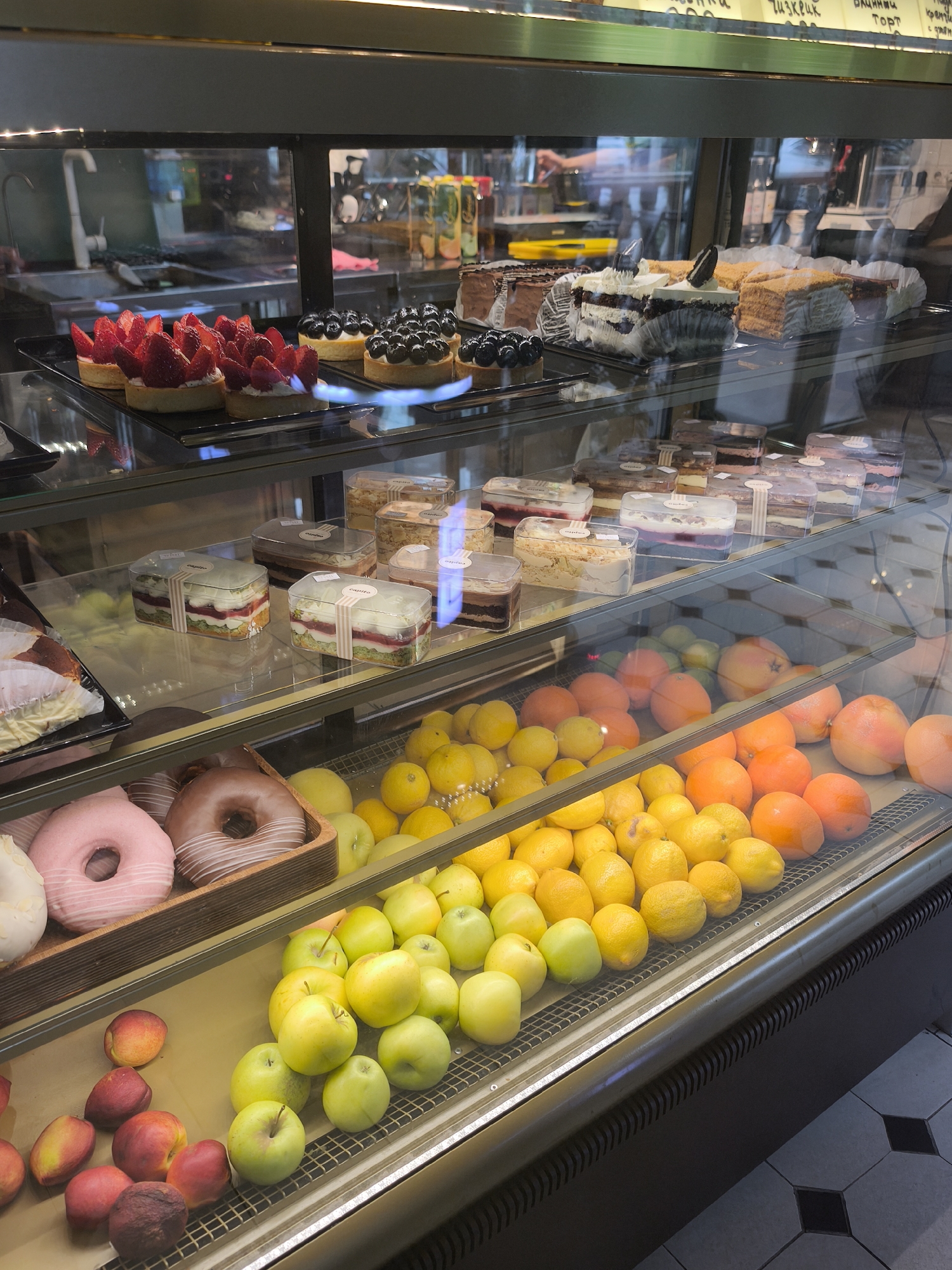The image shows a display case in a bakery or pastry shop in an unknown city. The case has multiple tiers and is filled with an assortment of desserts and fresh fruit. The top shelf displays pastries such as strawberry tarts, blueberry tarts, chocolate cake, and layered cakes. The second shelf has packaged layered desserts in transparent containers. The third shelf contains donuts in a wooden tray, as well as piles of lemons and oranges. The bottom shelf has piles of green apples and peaches. The display case is well-lit. The background is blurred, but it appears to be a bakery interior with visible kitchen equipment and other customers. The floor in front of the case has a black and white tiled pattern.