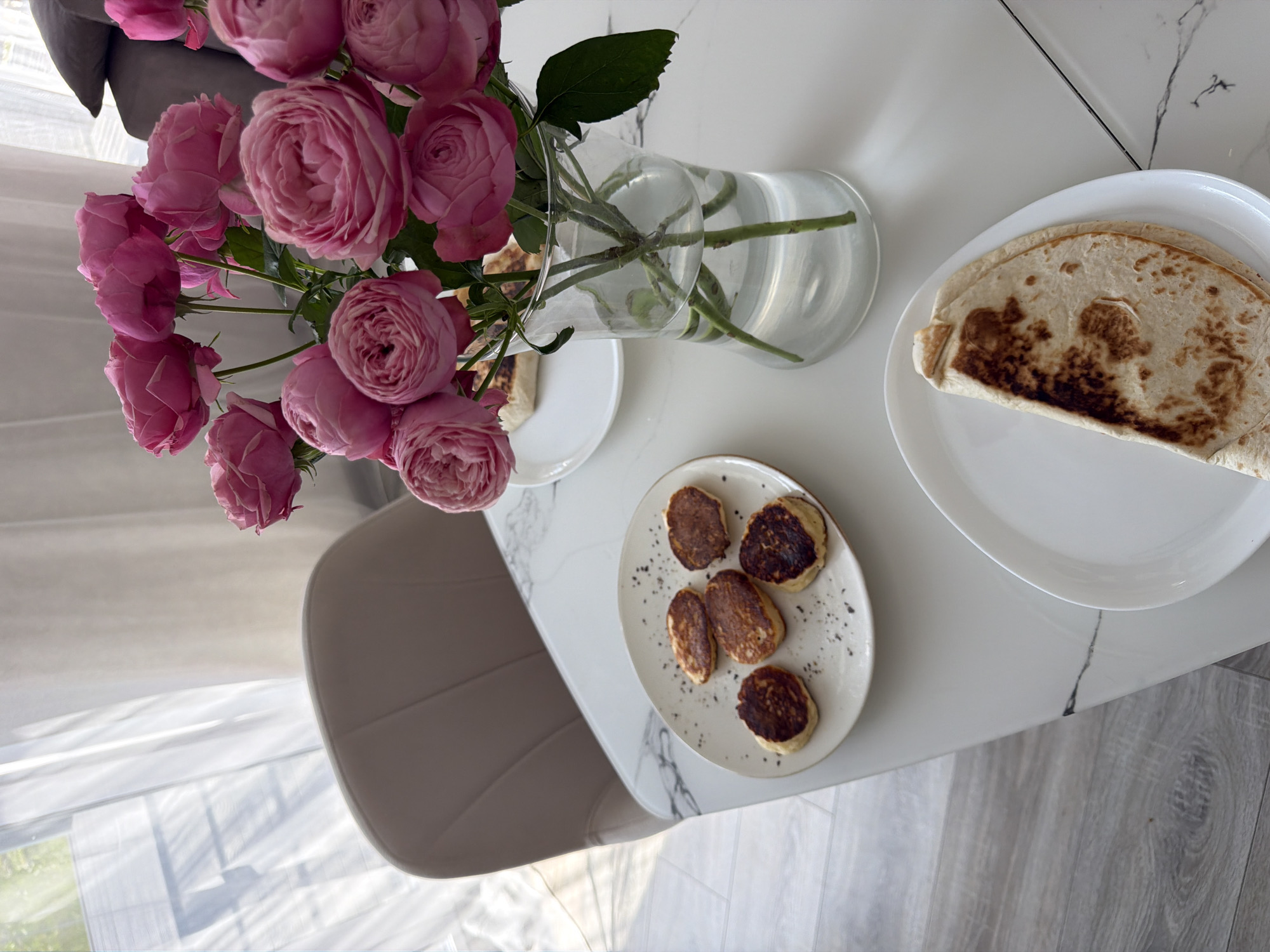 A table setting is depicted, featuring a vase of pink roses, a plate of small pancakes, and a plate with a folded tortilla. The table has a white surface with a black marble pattern and is situated near a window with sheer white curtains. In the background, a portion of a beige chair is visible. The scene suggests a meal or a decorative arrangement in what appears to be a domestic setting. There is no indication of the city of Malinovka, Russia, or any forensic details.
