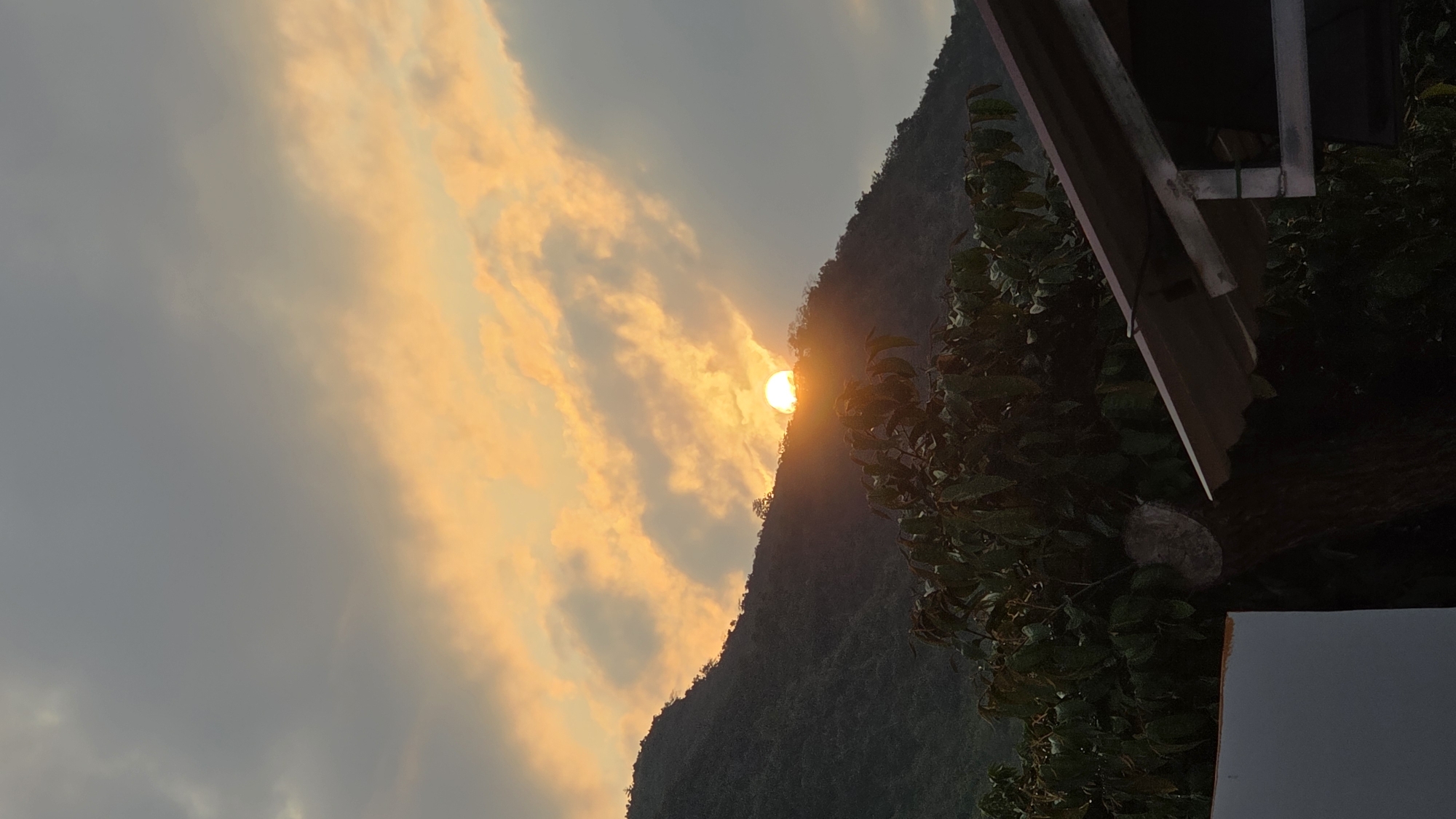 The image shows a mountain partially obscuring the setting sun in Bac Natural Heritage Area Trang, Vietnam. The sun casts an orange glow on the clouds and the sky, and it shines behind a mountain covered in trees. A building with an overhanging roof is visible in the right corner of the image.
