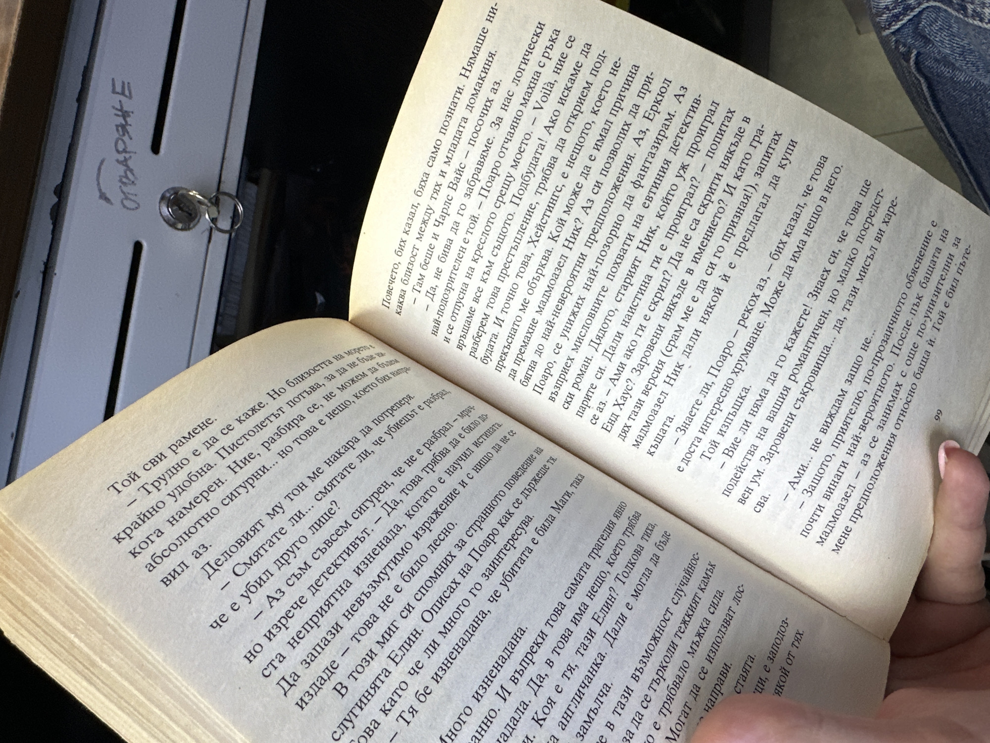 Here's a detailed description of the image:
**Scene:** The image shows an open book being held, likely in someone's lap, with the pages visible to the camera. The book is in Bulgarian, based on the Cyrillic script of the text. The page numbers are visible, with one page showing "99". The pages are aged, with a yellowed tone.
**Objects:**
* Book: The central object, showing text in Bulgarian.
* Hands: The hands holding the book.
* Metallic Surface: The upper left corner has a metallic surface with a keyhole and the Bulgarian word "ОТВАРЯНЕ" written above it, which translates to "OPENING".
**Environment:** The background is slightly out of focus, but it seems the book is likely being held indoors. The metal surface suggests it's near some kind of furniture or a cabinet.
**Forensic Details:**
* The lighting is subdued, suggesting either indoor lighting or a cloudy day.
* The focus of the image is sharpest on the book's pages, indicating that's the intended subject.
* The texture of the book's pages appears slightly rough.
**Location (Inferential):** Given the Bulgarian text, it can be inferred that the location is likely in Bulgaria. Without additional information, pinpointing a specific location in Sofia is not possible.