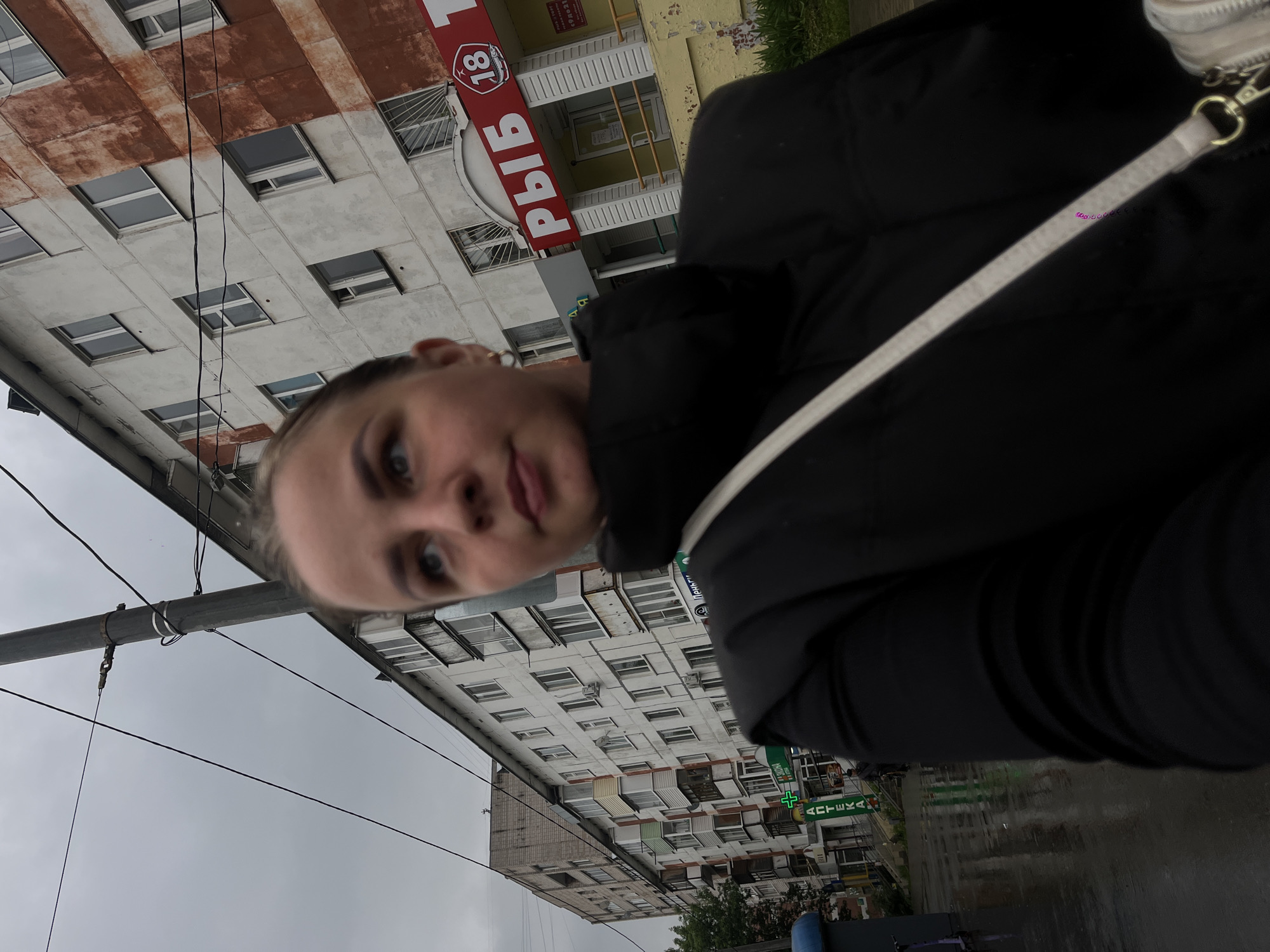 A medium shot depicts a woman wearing a black jacket and white purse strap, with a building in the background. The building has the sign "РЫБ" ("Fish" in Russian) and the number 18. The sign "АПТЕКА" ("Pharmacy" in Russian) is also visible. This suggests the location is Izhevsk, Russia, as the language on the signs is Russian. The sky is overcast, and the street appears wet, indicating recent rain.