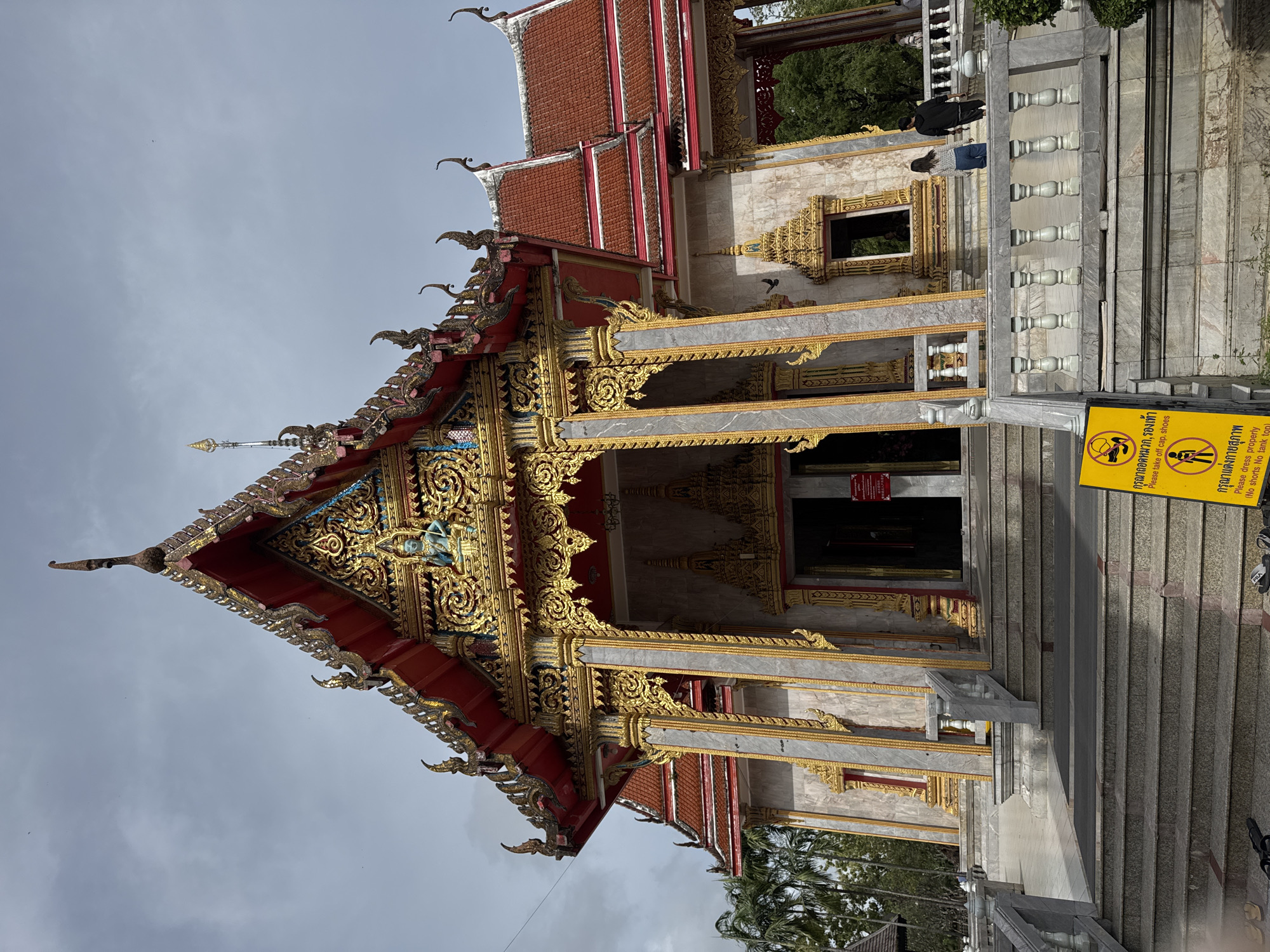 The image depicts the facade of a Thai Buddhist temple in Chalong, Thailand, under an overcast sky. The temple features ornate, multi-tiered roofs with red tiles and elaborate gold trim. The central entrance is flanked by tall, decorated pillars and leads into a dark interior. A statue of a seated figure, possibly a deity or Buddha, is visible on the upper part of the structure.
In front of the temple is a wide set of stairs leading up to the entrance, with grey marble balustrades on either side. A yellow sign with black symbols and Thai text is prominently placed at the foot of the stairs, indicating rules for visitors, including a prohibition against wearing a cap and shoes, and a requirement to dress properly, excluding tank tops. Two individuals are visible on the temple's upper level, one in a black hoodie and the other in a white top and blue jeans, facing away from the camera and towards the temple entrance. Another person is partially visible further to the right. Palm trees are visible on the left side of the image, and lush green trees line the background on the right. The overall impression is a solemn and culturally significant architectural site.