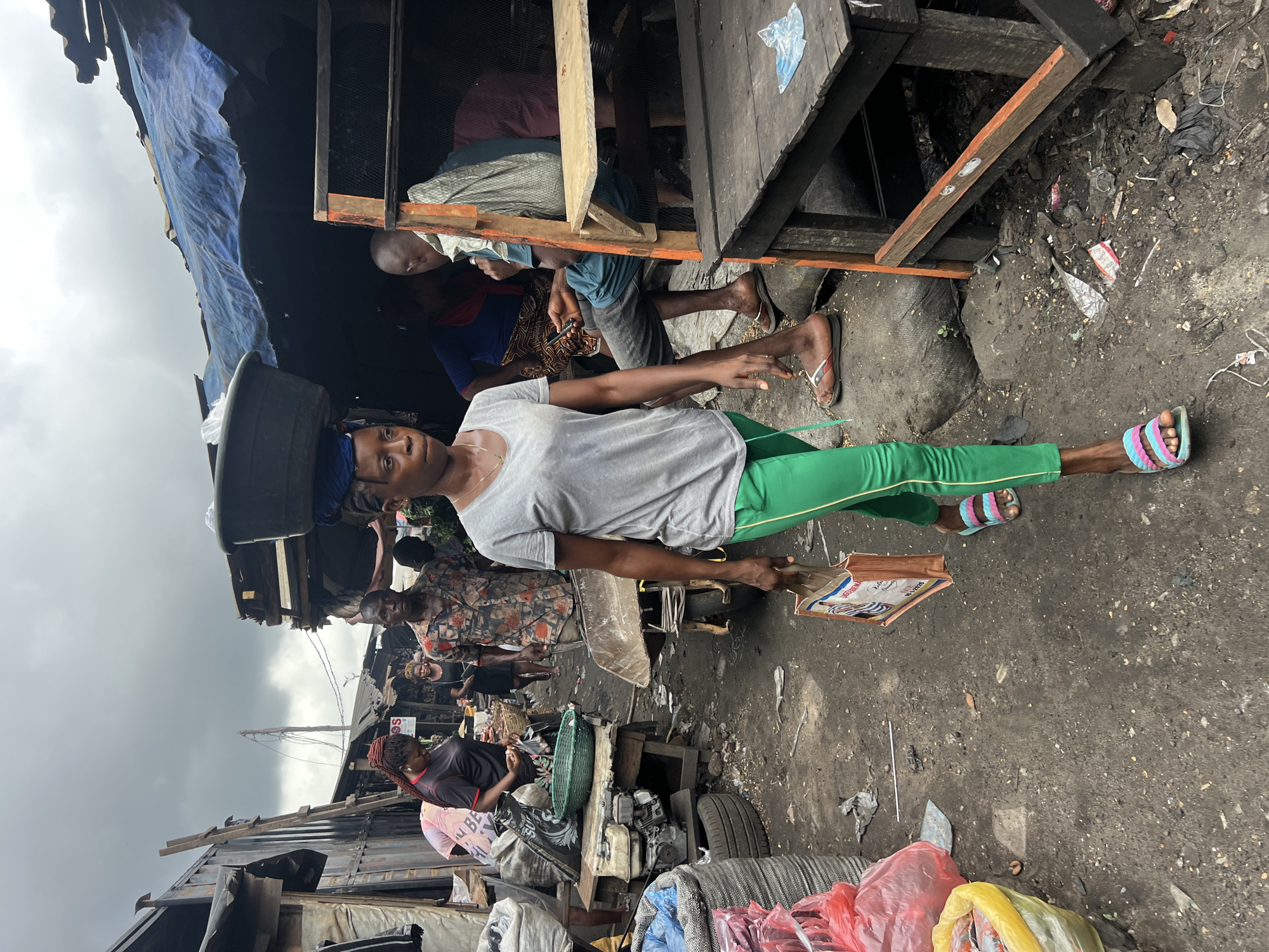 A medium shot depicts a woman carrying a container on her head and a cardboard container in her right hand. She is wearing a gray t-shirt and green leggings. The location appears to be a street market in the city of Unknown, Unknown. People, buildings, and debris are present in the background.