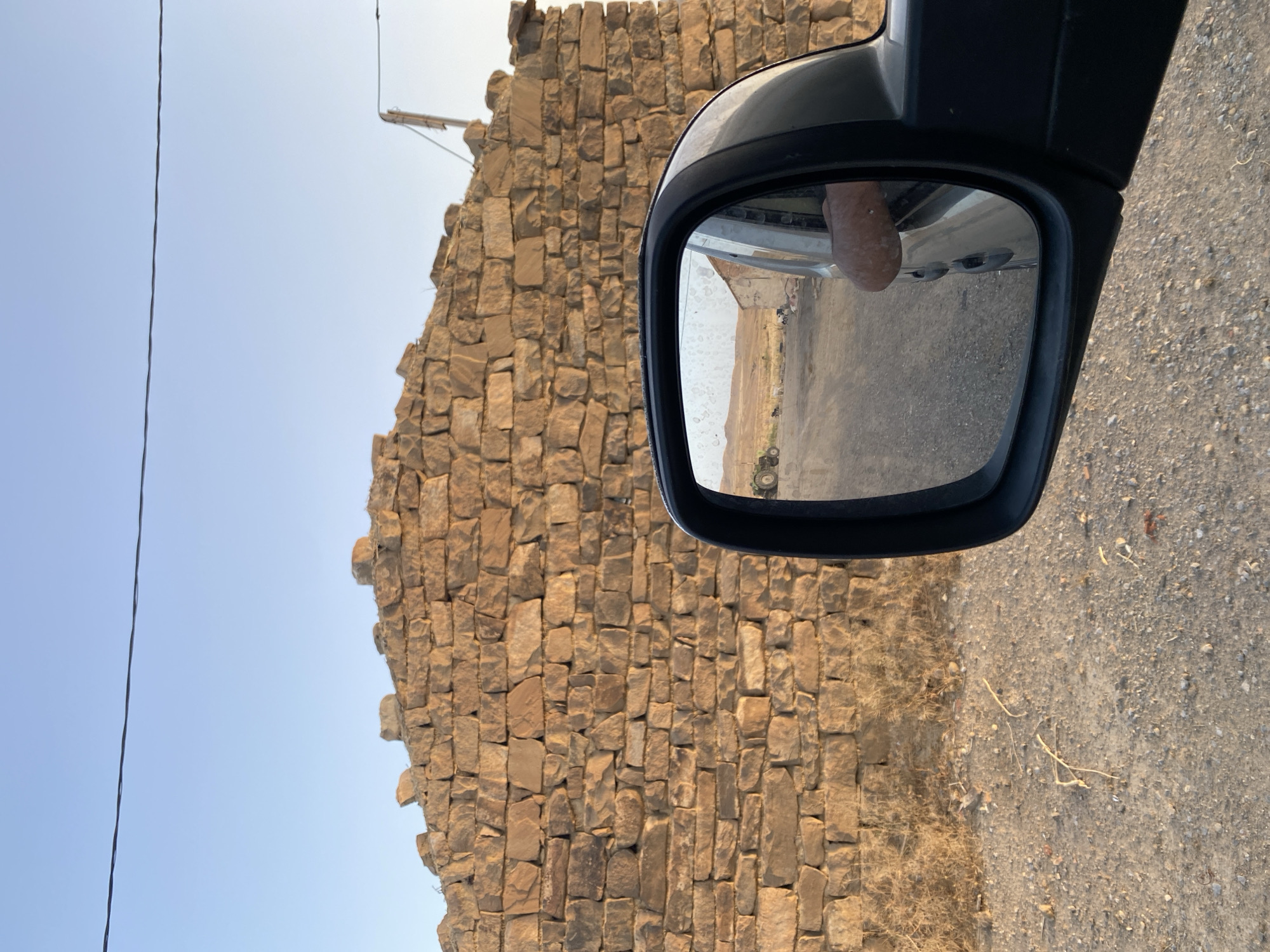 The image depicts a rural scene captured from a vehicle, with a significant portion of the frame dominated by a weathered, ancient stone wall constructed from large, irregular blocks. The wall appears to be a ruin, with a rough, uneven top edge. To the right of the wall, a segment of a modern vehicle's side mirror is visible, reflecting a dusty road, a distant building, and a person's arm resting on the car window. In the reflection of the mirror, a green tractor is parked to the left. The ground in the foreground is dirt and gravel, with sparse, dry vegetation. The sky above is clear and a pale blue, suggesting a warm, dry climate. A single black wire stretches horizontally across the upper part of the image, indicating the presence of electrical infrastructure. While the general setting is clearly rural and appears to be in North Africa, the specific location within the city of Aziz, Algeria, cannot be confirmed from the visual information provided.