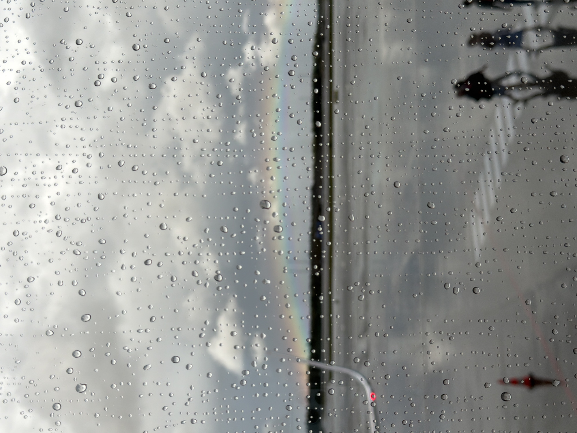 This image, taken in Gardermoen, Norway, through a window covered in raindrops, depicts a somewhat overcast sky with a faint rainbow visible in the distance. The foreground is dominated by the water droplets on the glass, creating a blurred effect on the scene outside.
The exterior environment appears to be an airport tarmac. In the mid-ground, there is a faint, horizontal line of dark green foliage or trees. Beyond this, the tarmac is wet, reflecting the gloomy sky. Several out-of-focus figures are walking across the tarmac in the right-middle ground, their forms indistinct due to the rain and distance. Some appear to be carrying luggage. Further to the left, the wing of an aircraft is visible, with a red navigation light illuminated. A red and white traffic cone is also present on the tarmac in the lower-left quadrant. The overall impression is of an airport on a rainy day, with the natural phenomenon of a rainbow offering a subtle splash of color against the muted tones.