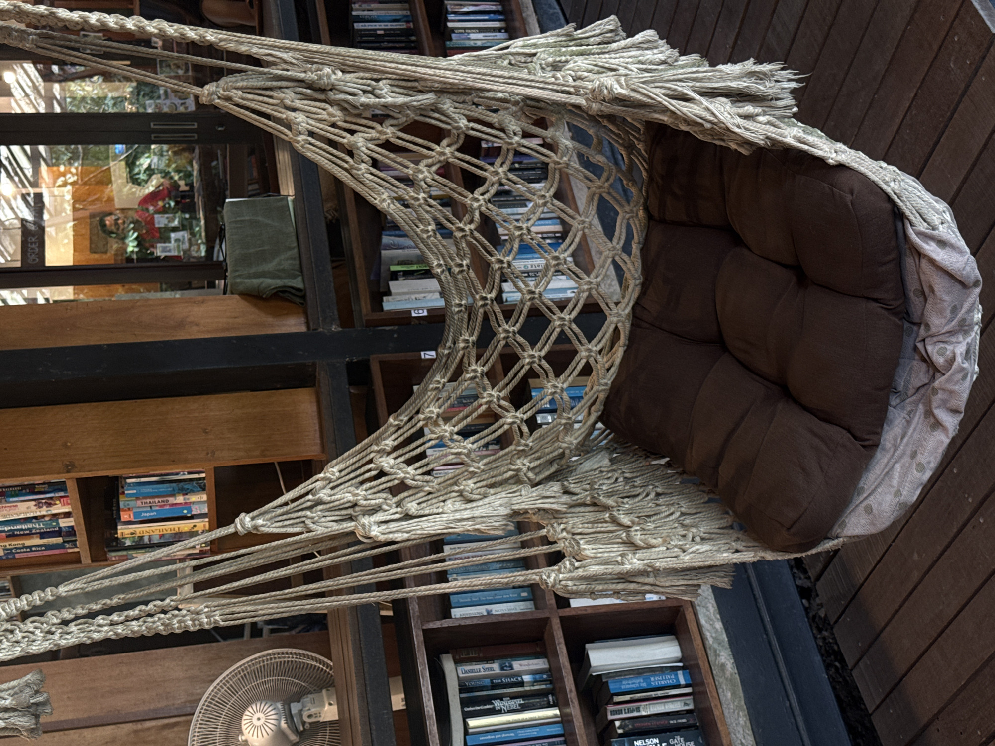 A macrame hammock chair, filled with a tufted brown cushion, is suspended in front of bookshelves and a window. The hammock is constructed of off-white rope, with braided supports and tassels. The cushion is a dark brown, deeply tufted, and rests on a patterned, light-colored fabric base. Behind the hammock, wooden bookshelves are filled with books, some of which are visible on the left side of the image. To the right, a large window shows a blurred reflection of an indoor space with greenery and a person wearing a red shirt. The floor is made of dark wooden planks. A white fan is visible on a shelf to the left of the hammock. This scene appears to be set in a cafe or library, possibly located in Pa Khlok, Thailand, given the presence of a fan and the outdoor lighting visible through the window.